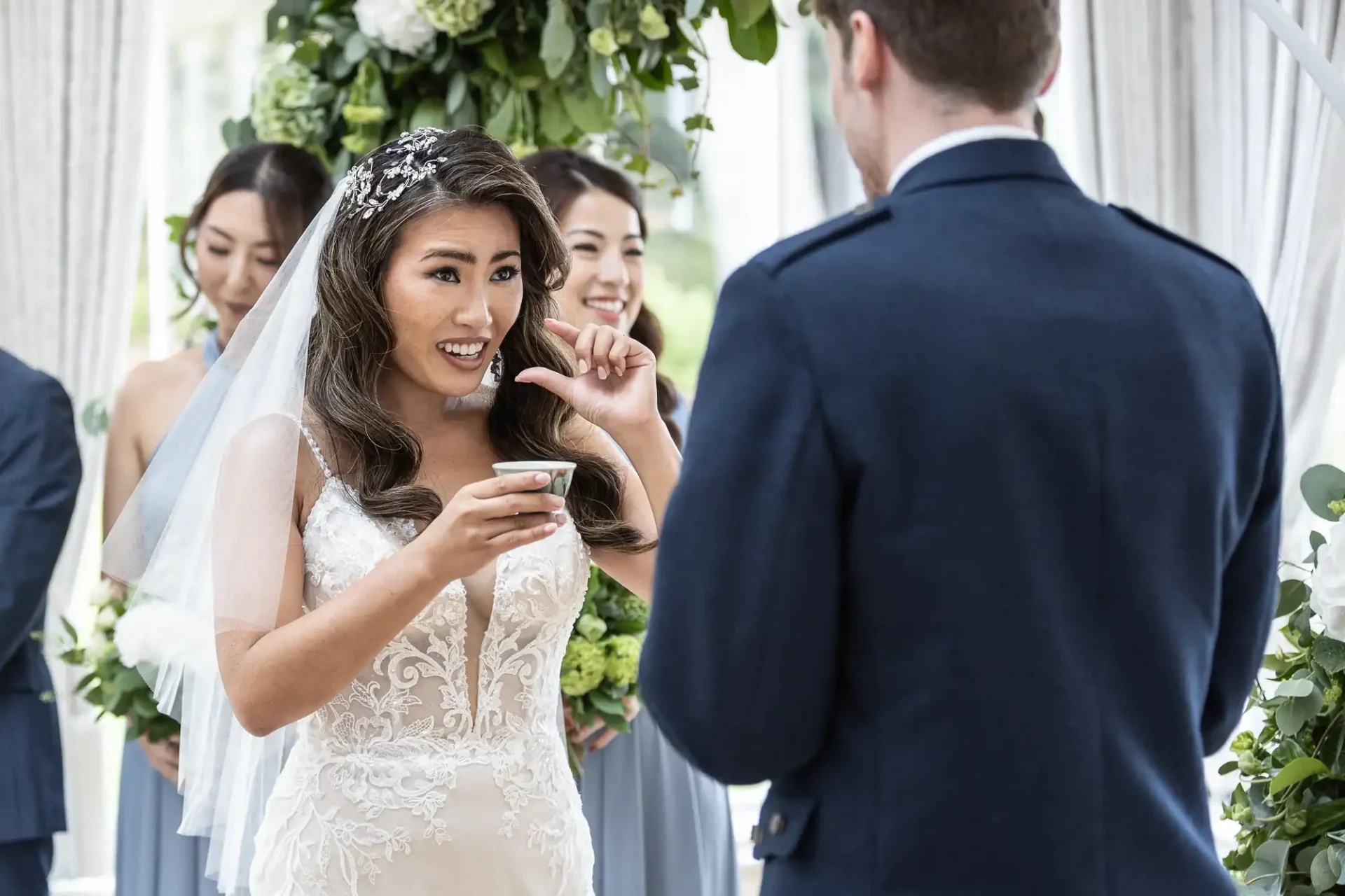 Bride holding a cup, gesturing with her hand, and smiling at the groom during a wedding ceremony. Bridesmaids in matching dresses stand behind her.