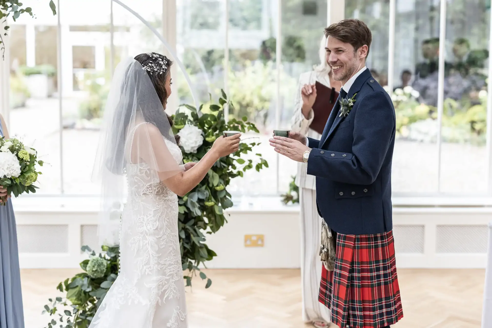 A bride and groom exchange cups during a wedding ceremony indoors. The groom wears a kilt and jacket, and the bride is in a white gown and veil. Greenery decorates the scene.