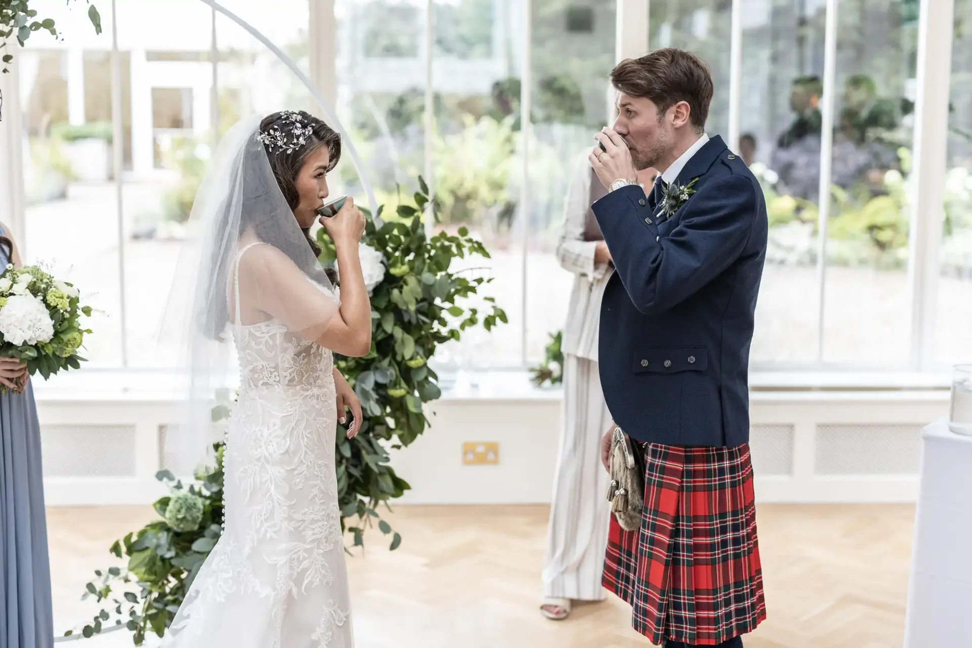 Bride and groom in a wedding ceremony, both drinking from cups. The groom is wearing a kilt, and the bride is in a lace gown with a veil. Greenery decor surrounds them.