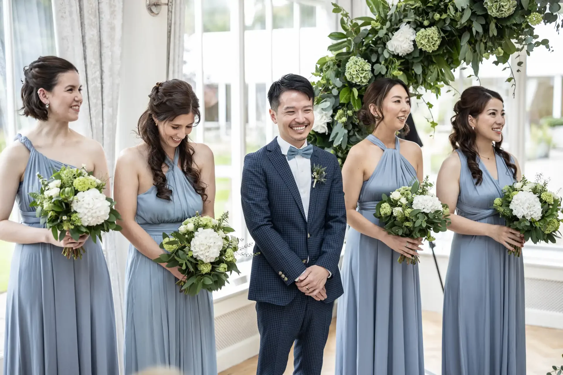 A groom in a blue suit stands with five bridesmaids in matching blue dresses, holding bouquets, in a bright room with a floral arch.