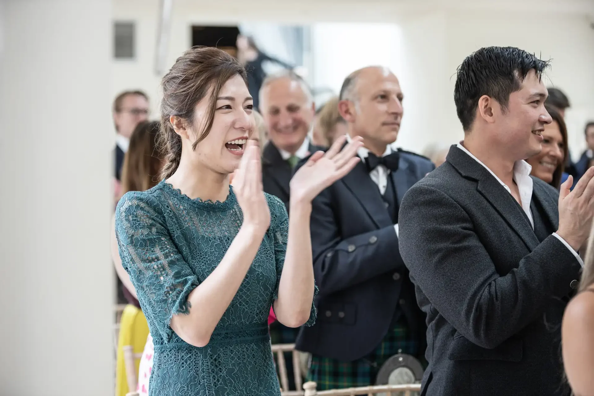 A group of people, including a woman in a green dress, clapping and smiling at an indoor event.