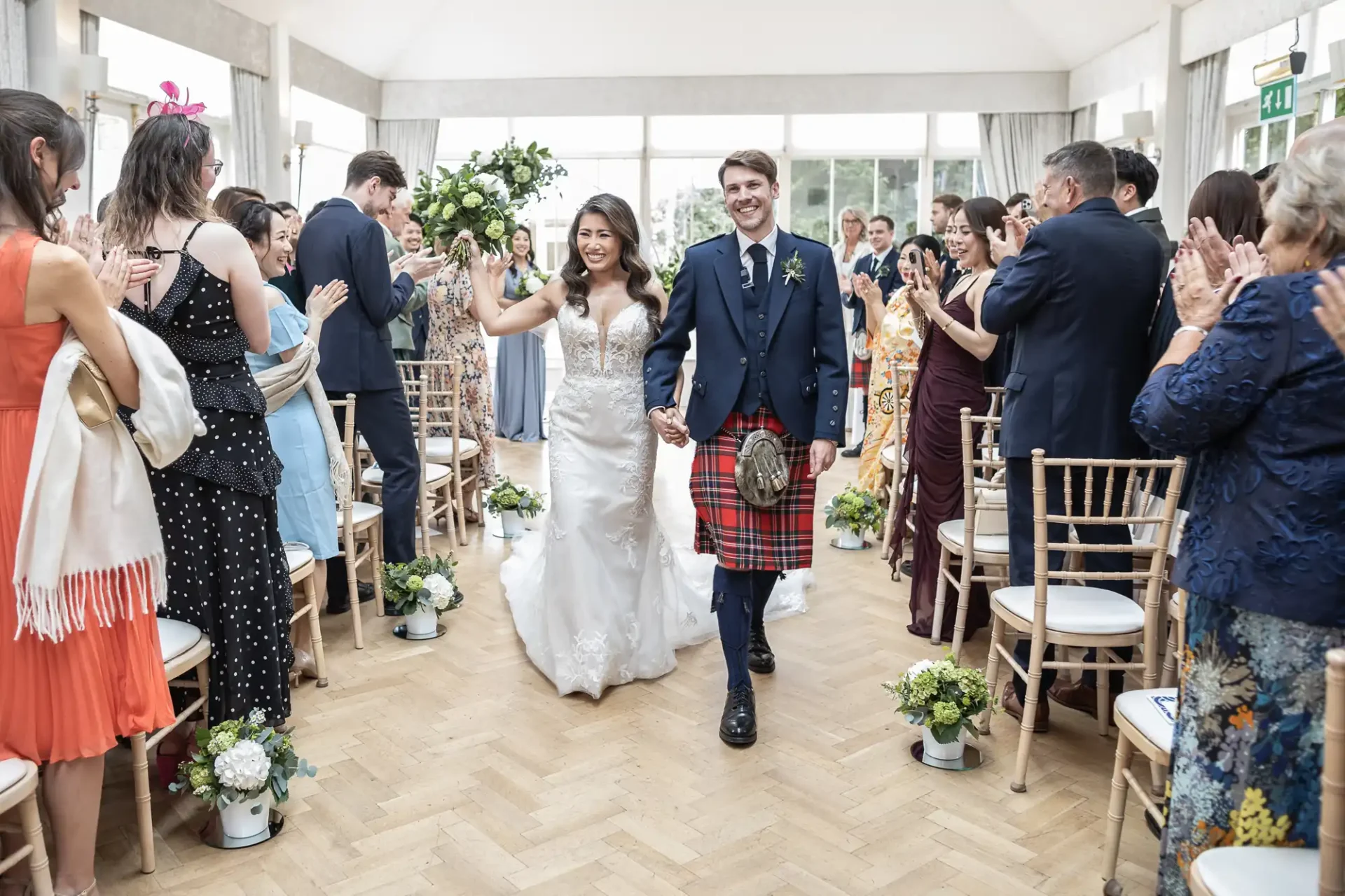 A couple in wedding attire walks down an aisle, surrounded by applauding guests in a bright room. The groom wears a kilt, and the bride holds a bouquet.