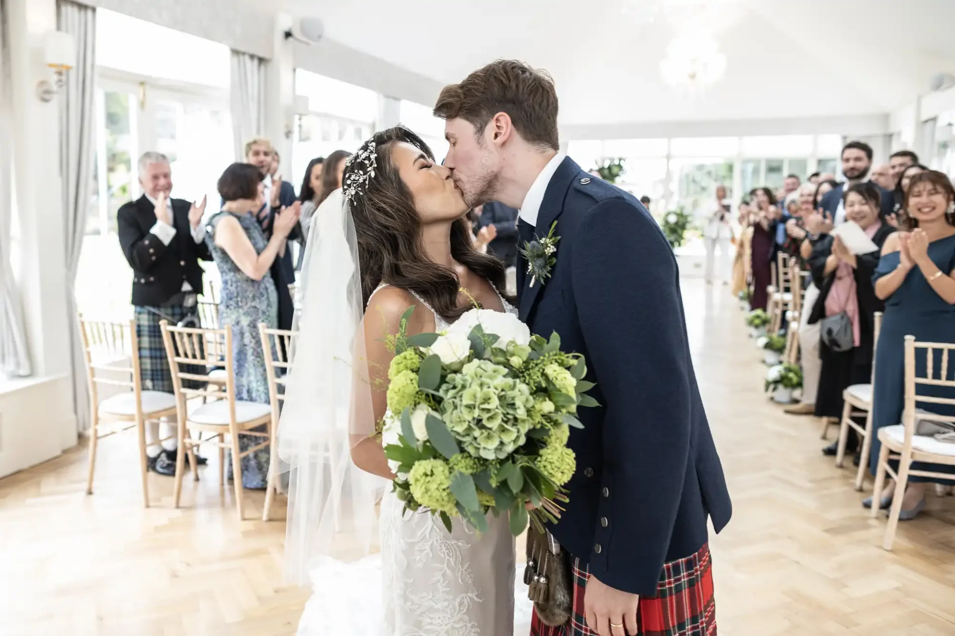 Bride and groom kiss at the end of the aisle in a bright room, surrounded by applauding guests. The groom wears a kilt, and the bride holds a bouquet.