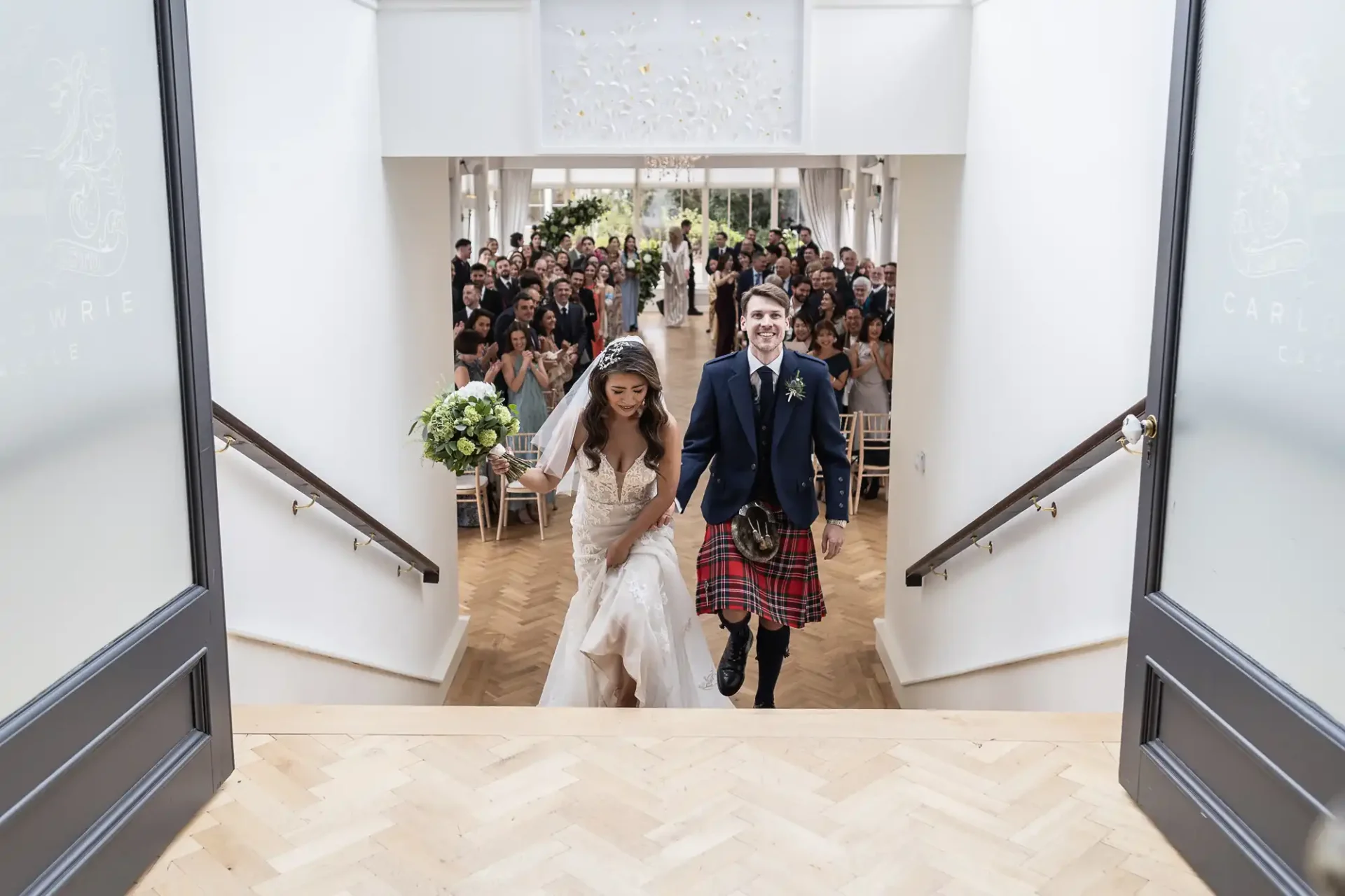 A bride and groom walk up stairs smiling, with guests seated behind them. The groom is wearing a kilt.