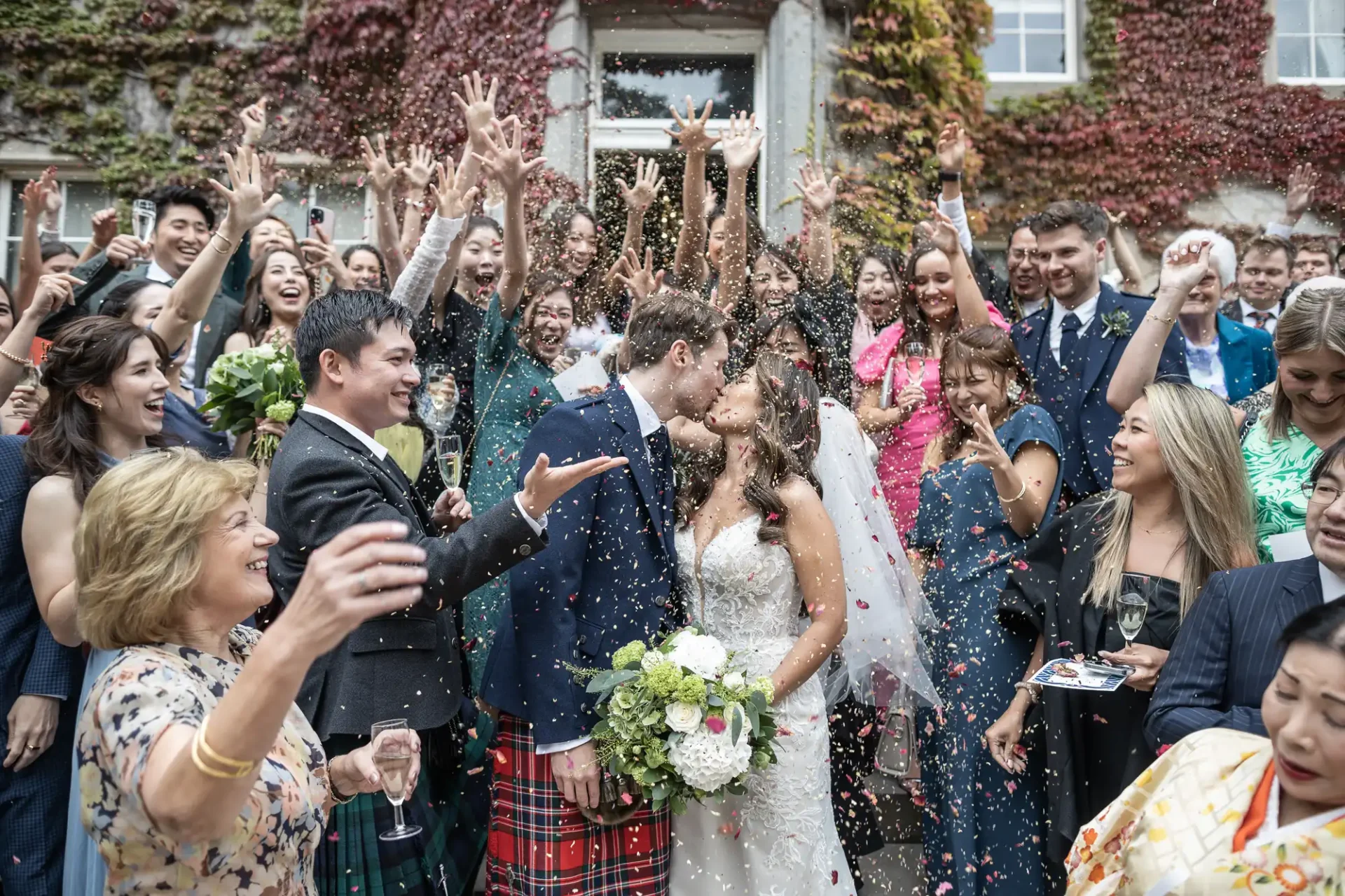 A couple kisses surrounded by cheering guests throwing confetti, with everyone dressed in formal attire and standing outside a building with ivy-covered walls.