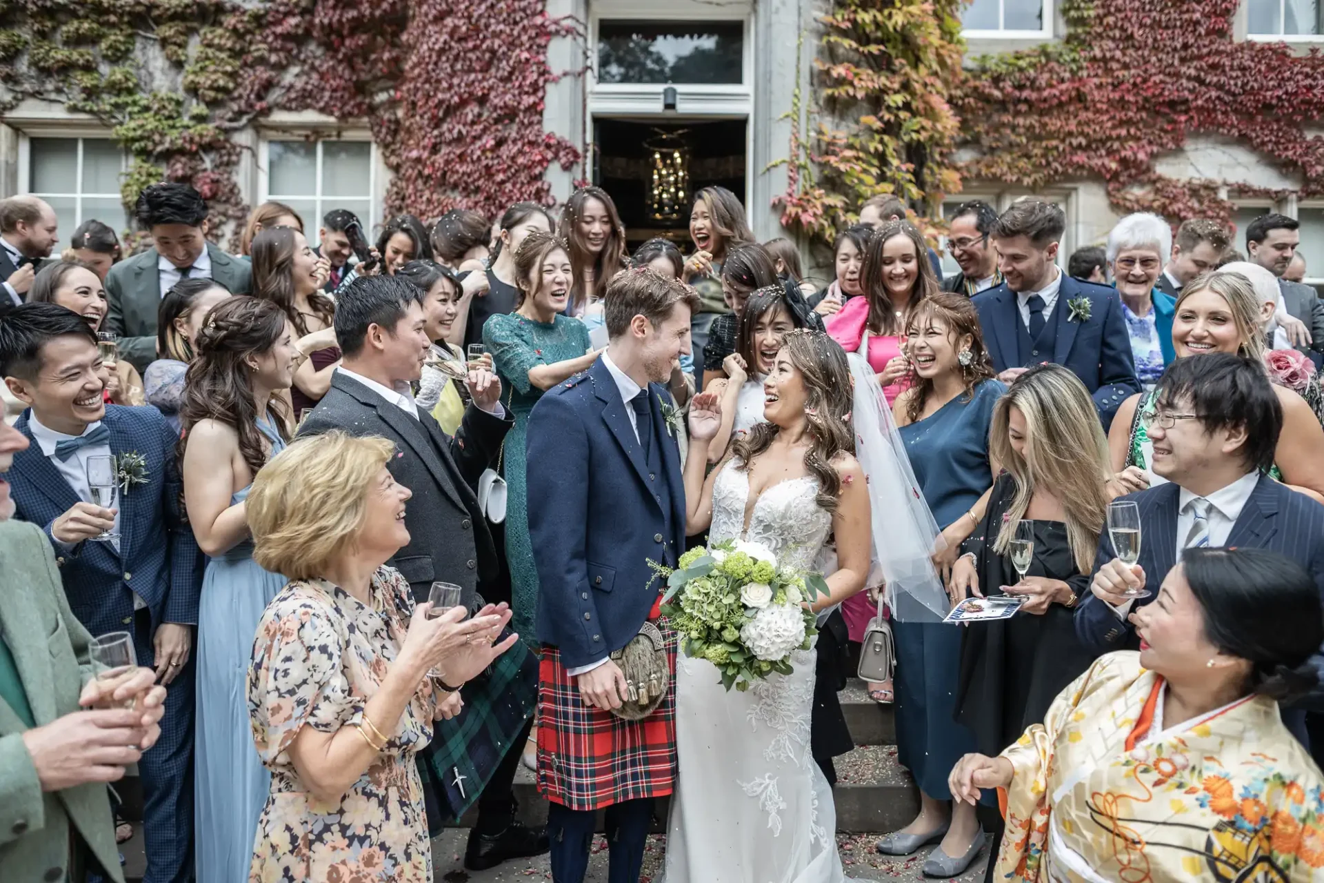 A newlywed couple stands smiling among a large group of guests outside, with some holding champagne glasses. The groom wears a kilt, and the bride holds a bouquet. Ivy-covered building in the background.