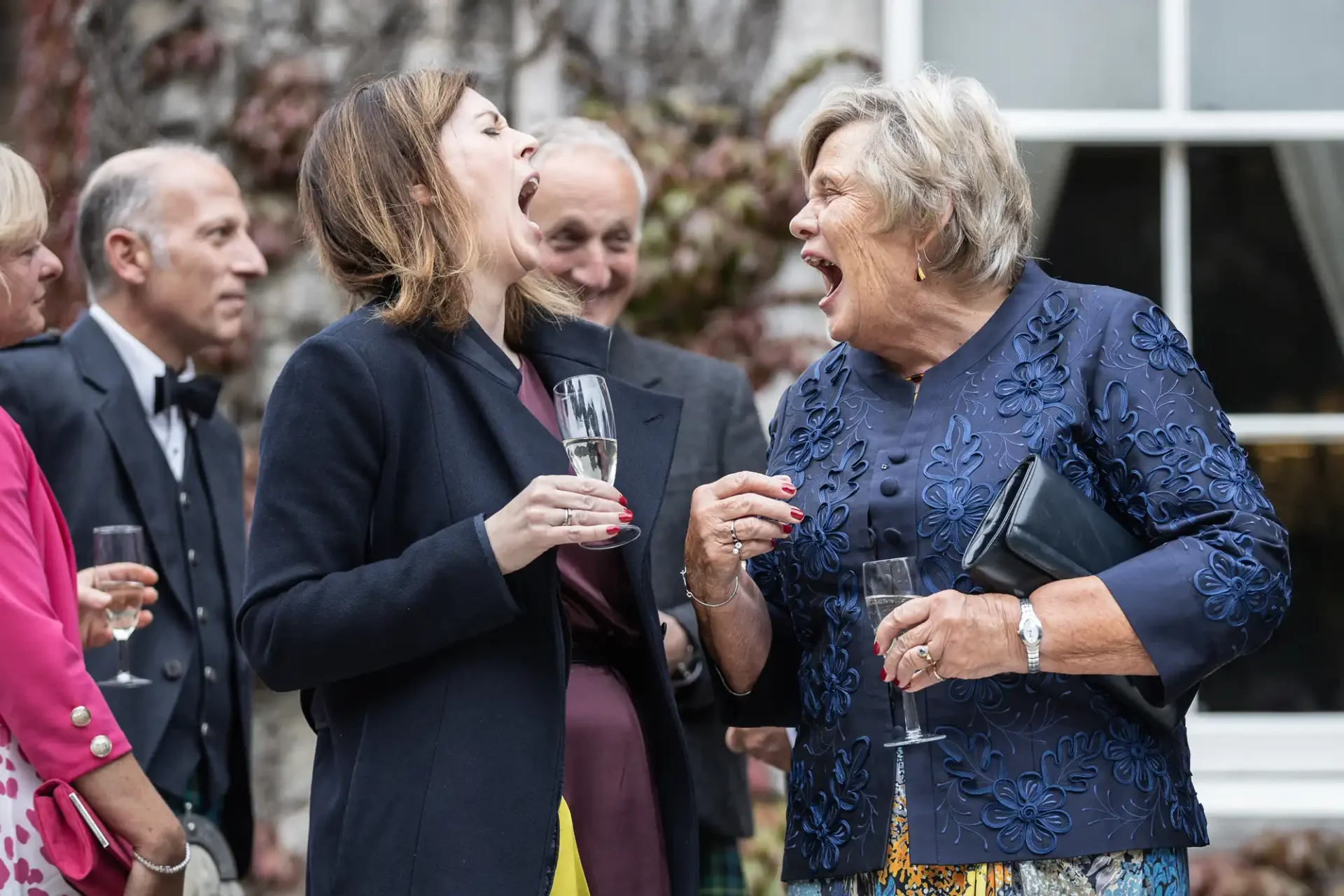 Two women stand outdoors laughing with open mouths, holding champagne glasses. People in formal attire are around them.