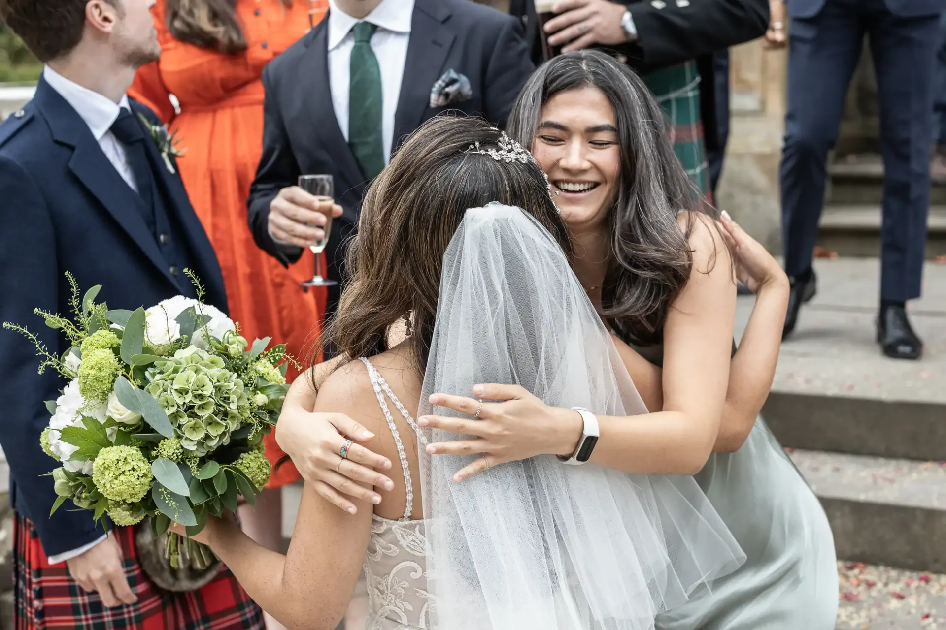 Bride holding a bouquet hugs a woman in a silver dress, surrounded by people dressed for a wedding.