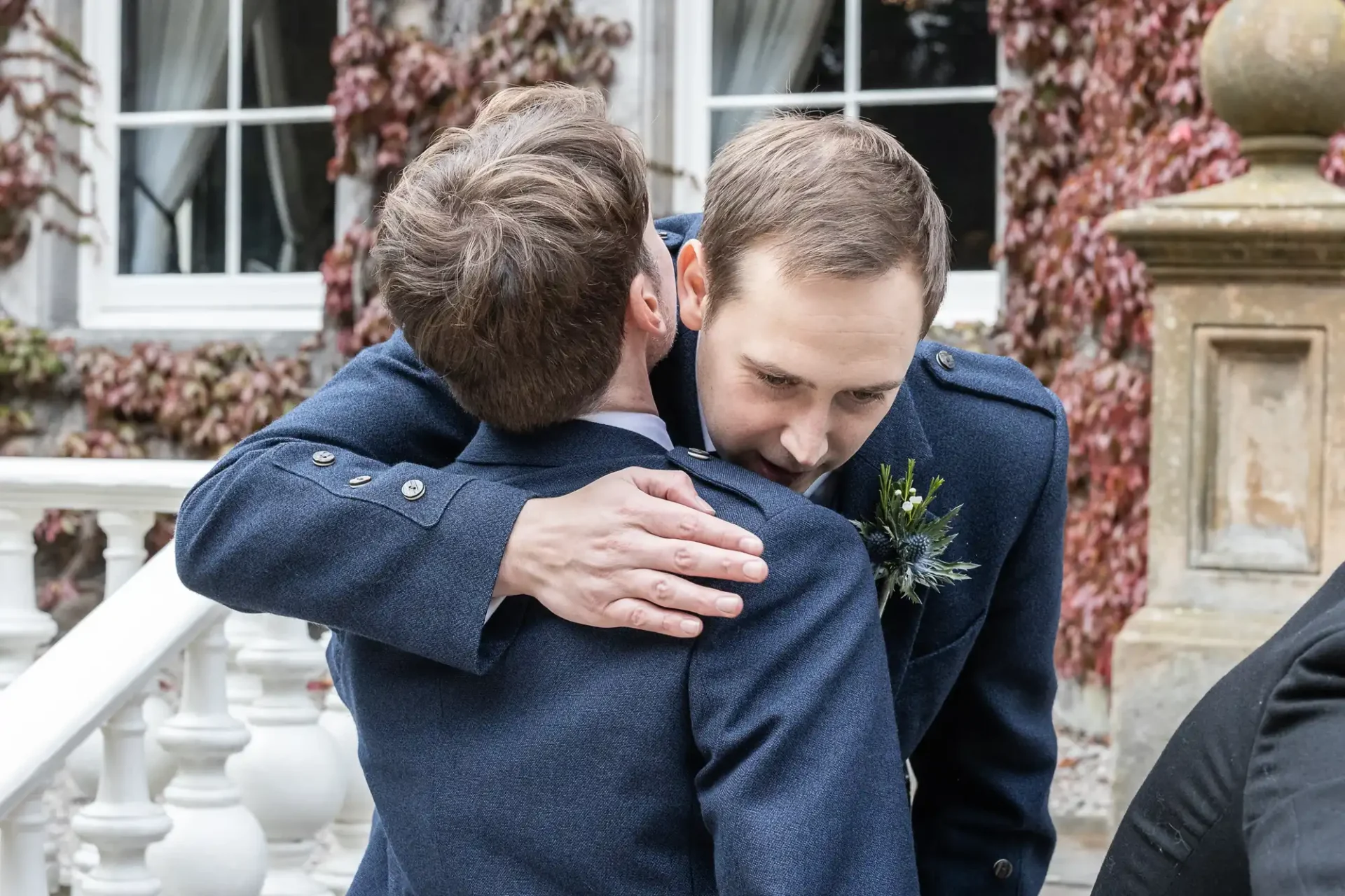 Two men in blue suits embrace near a balustrade, with ivy-covered building in the background.