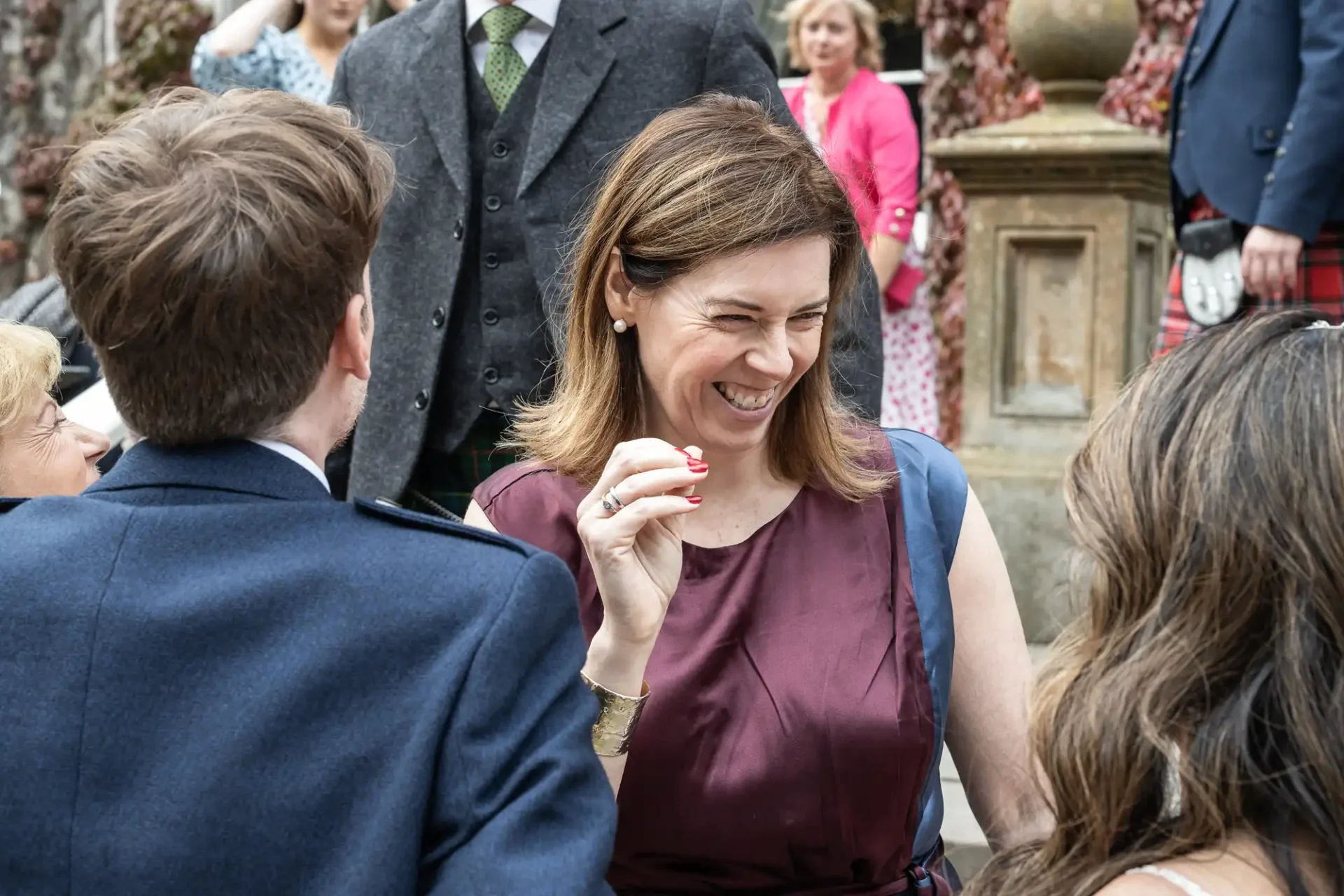 A woman in a burgundy dress smiles and interacts with others at an outdoor social event. People in formal attire surround her.