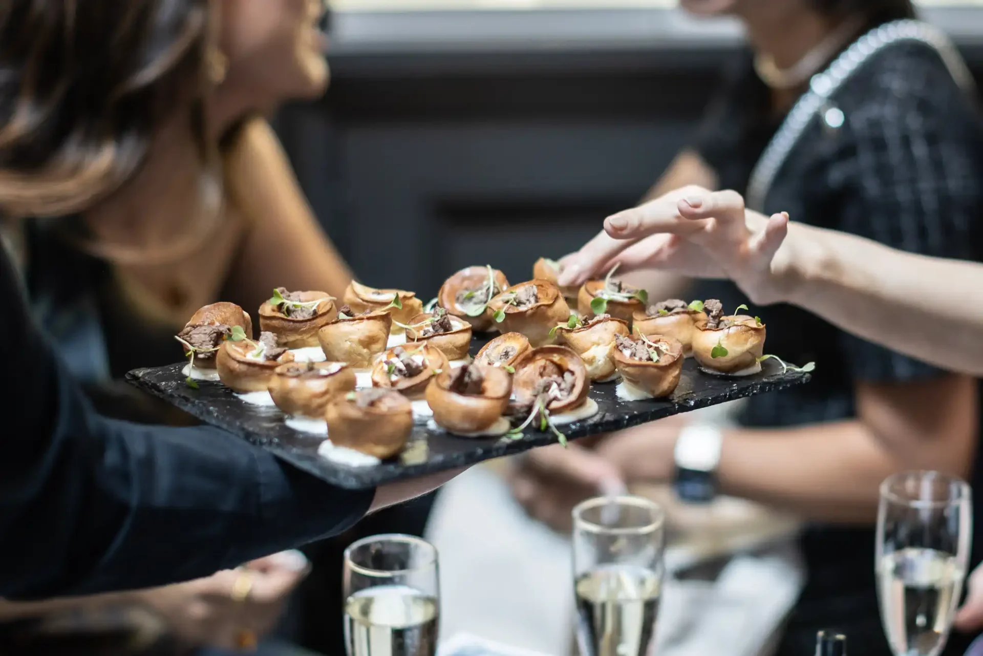 A tray of bite-sized appetizers being served to people. Three filled champagne glasses are on the table in front of them.