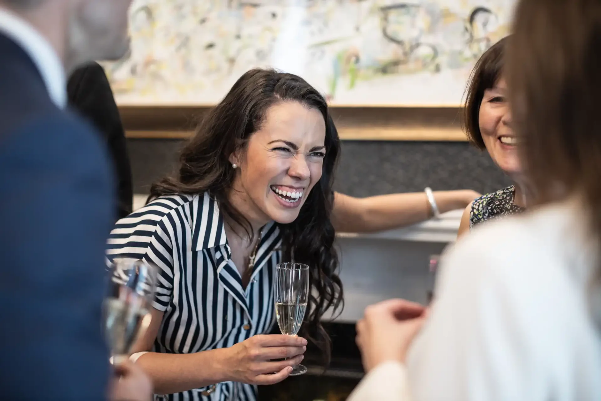 A woman in a striped dress is laughing while holding a glass at a social gathering with others.