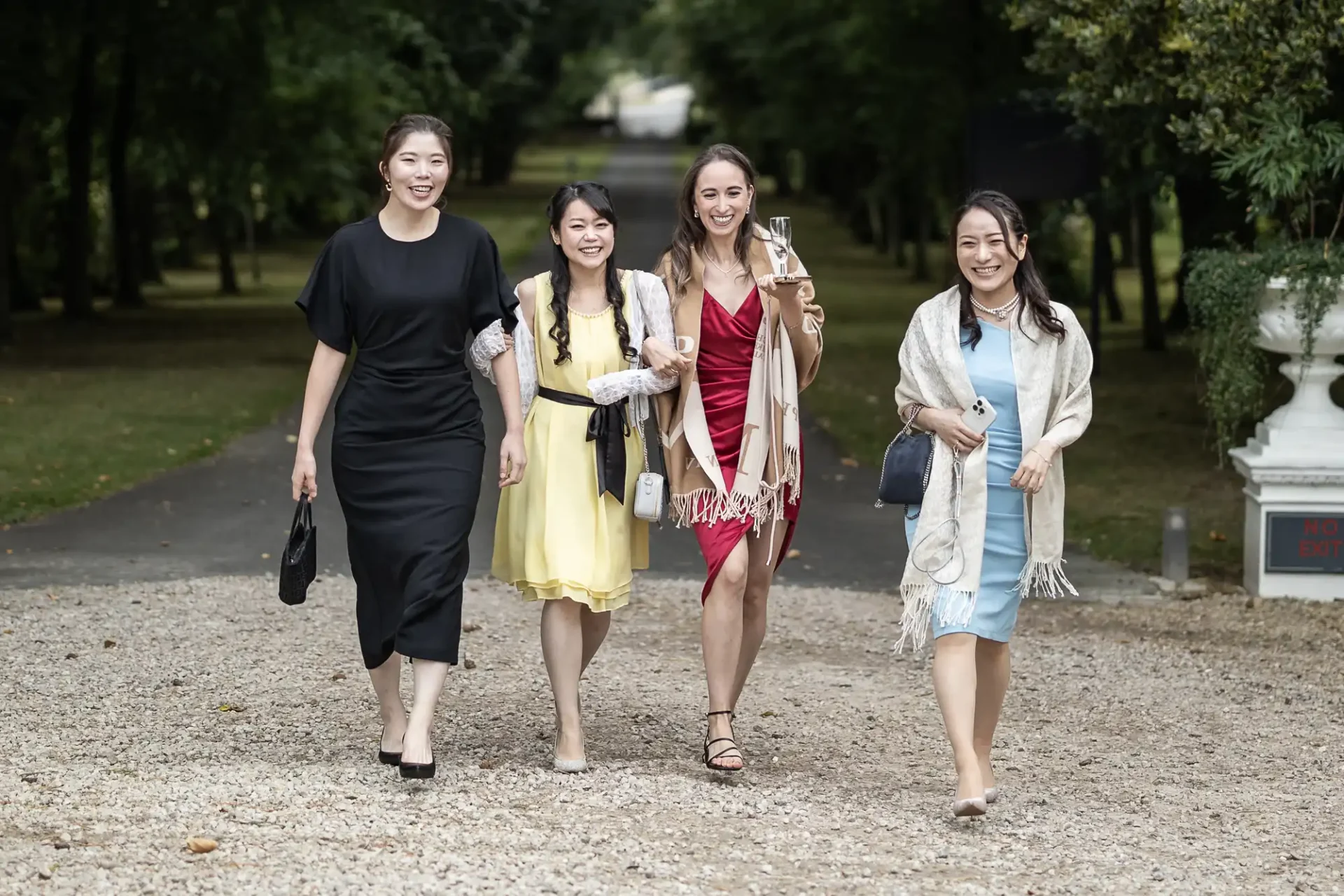 Four women walking outdoors on a gravel path, dressed in formal attire with one holding a wine glass, surrounded by trees.