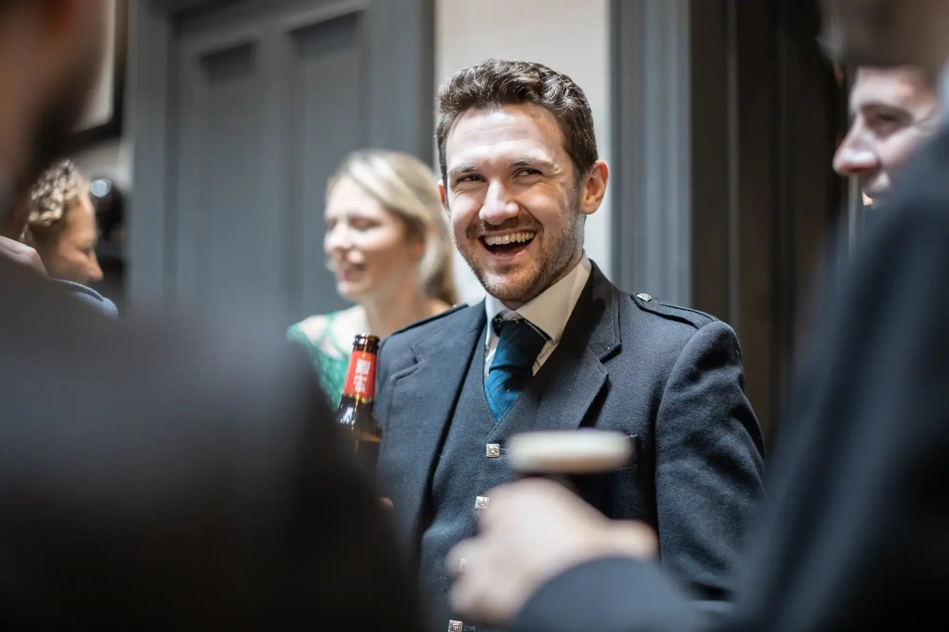 Man in formal attire holding a drink, smiling and standing with a group in a social setting.