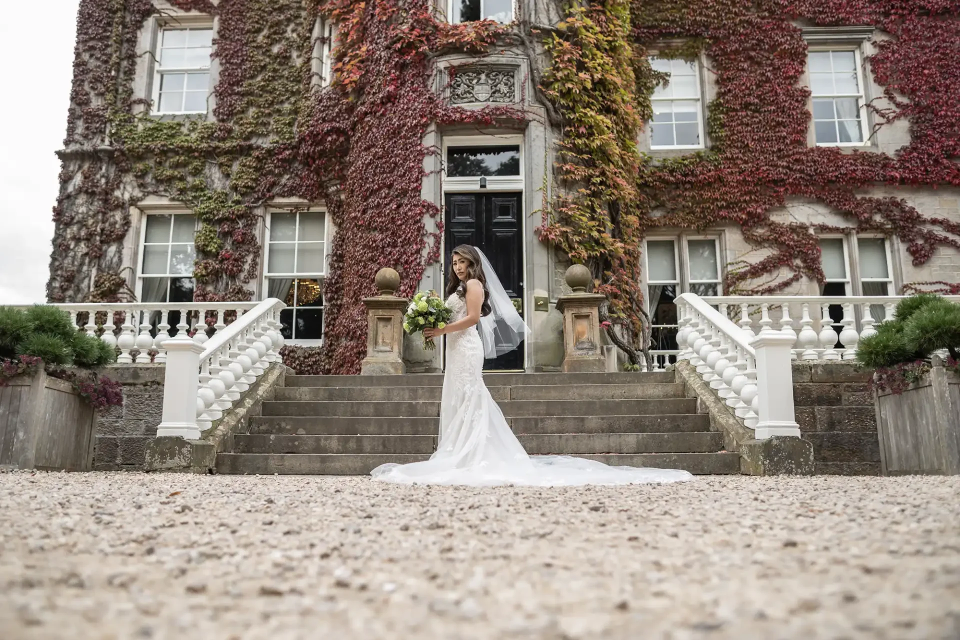 Bride in a white dress stands on steps in front of a large, ivy-covered building, holding a bouquet.