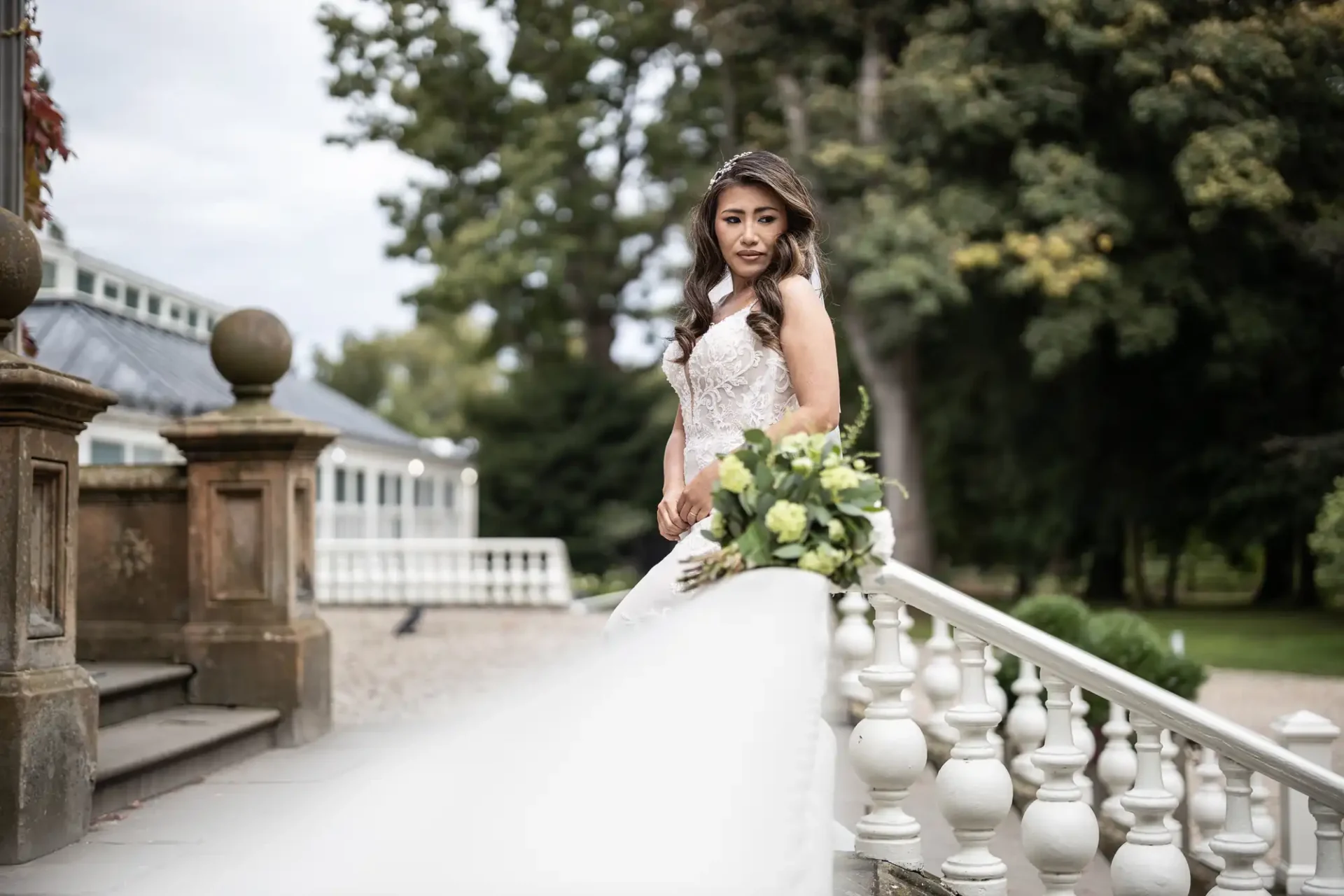 Bride in a white dress standing on stairs, holding a bouquet, with trees and a building in the background.