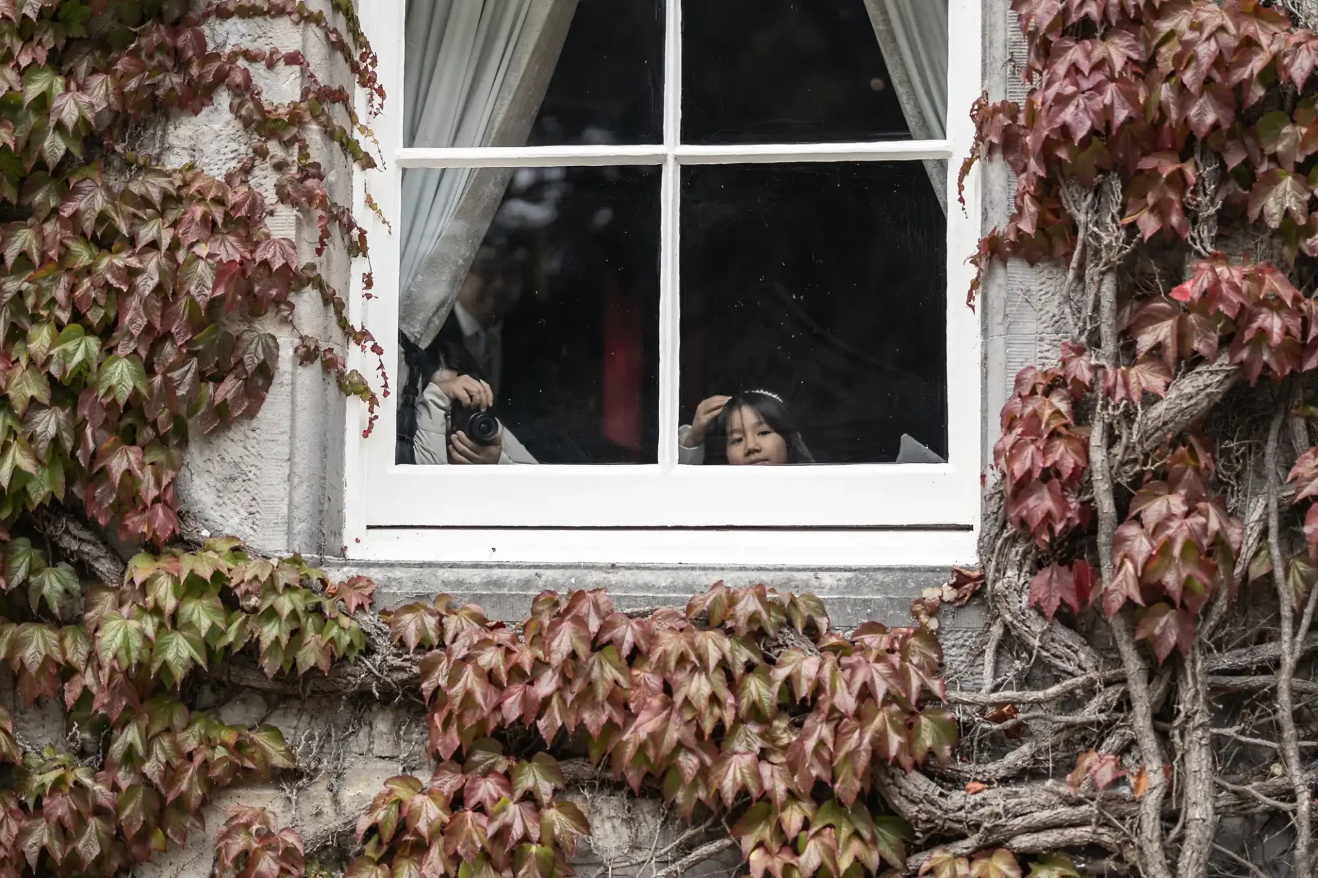 Two people are visible through a window framed by ivy-covered walls, one holding a camera.