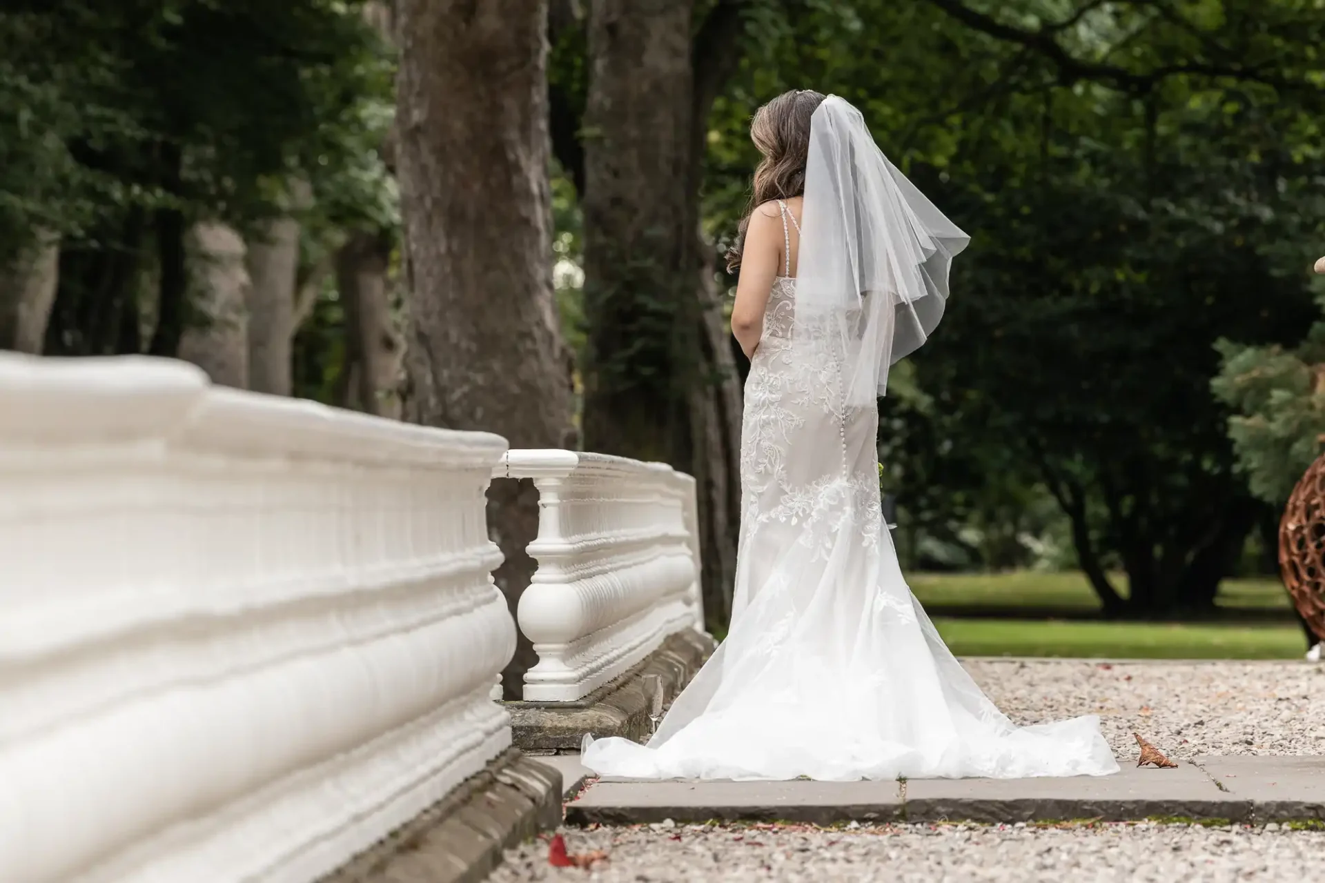 A bride in a white gown and veil stands outdoors near a white railing, facing away, amidst trees and greenery.