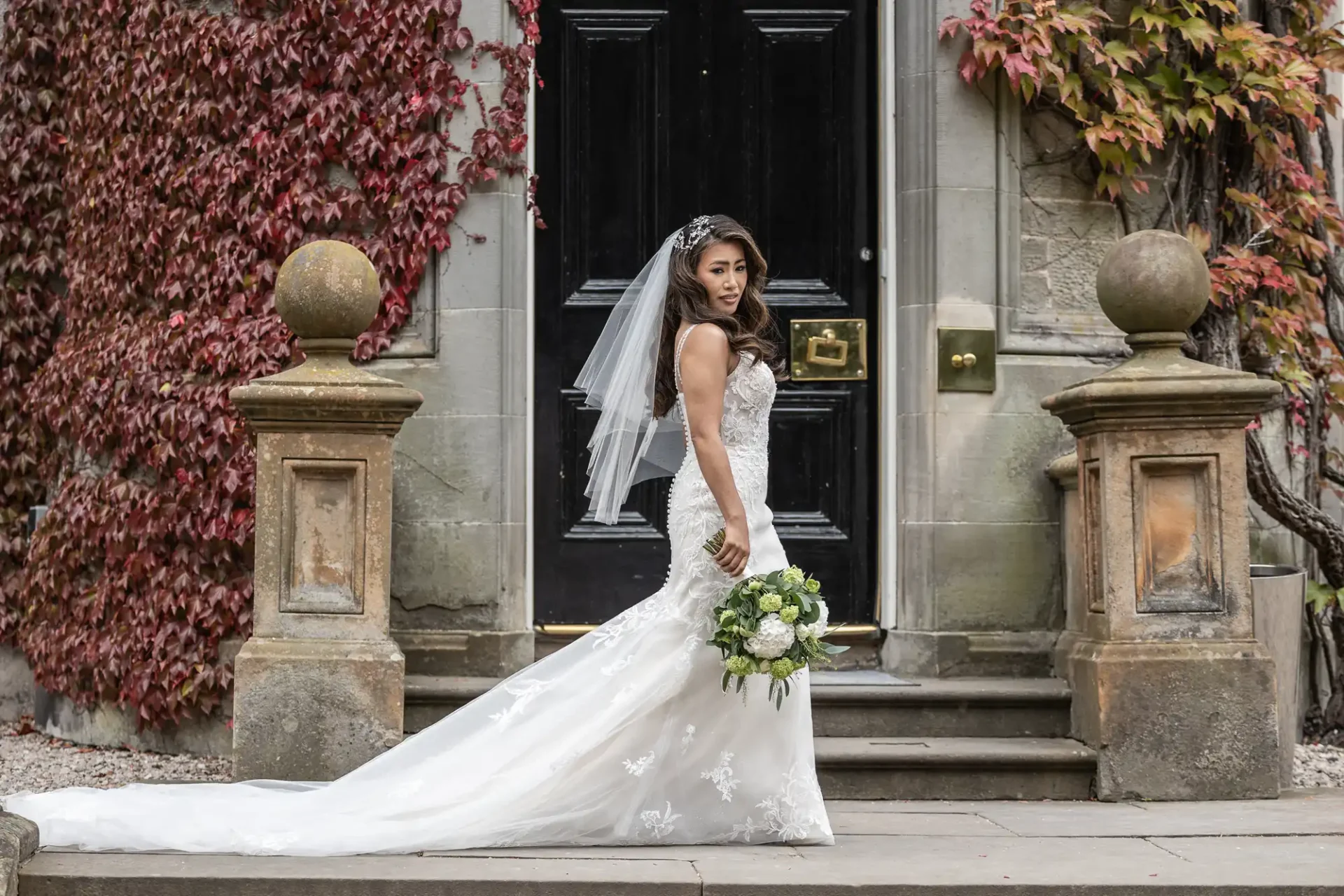 Bride in white wedding dress holding bouquet stands in front of a stately building with ivy-covered walls and a black door.