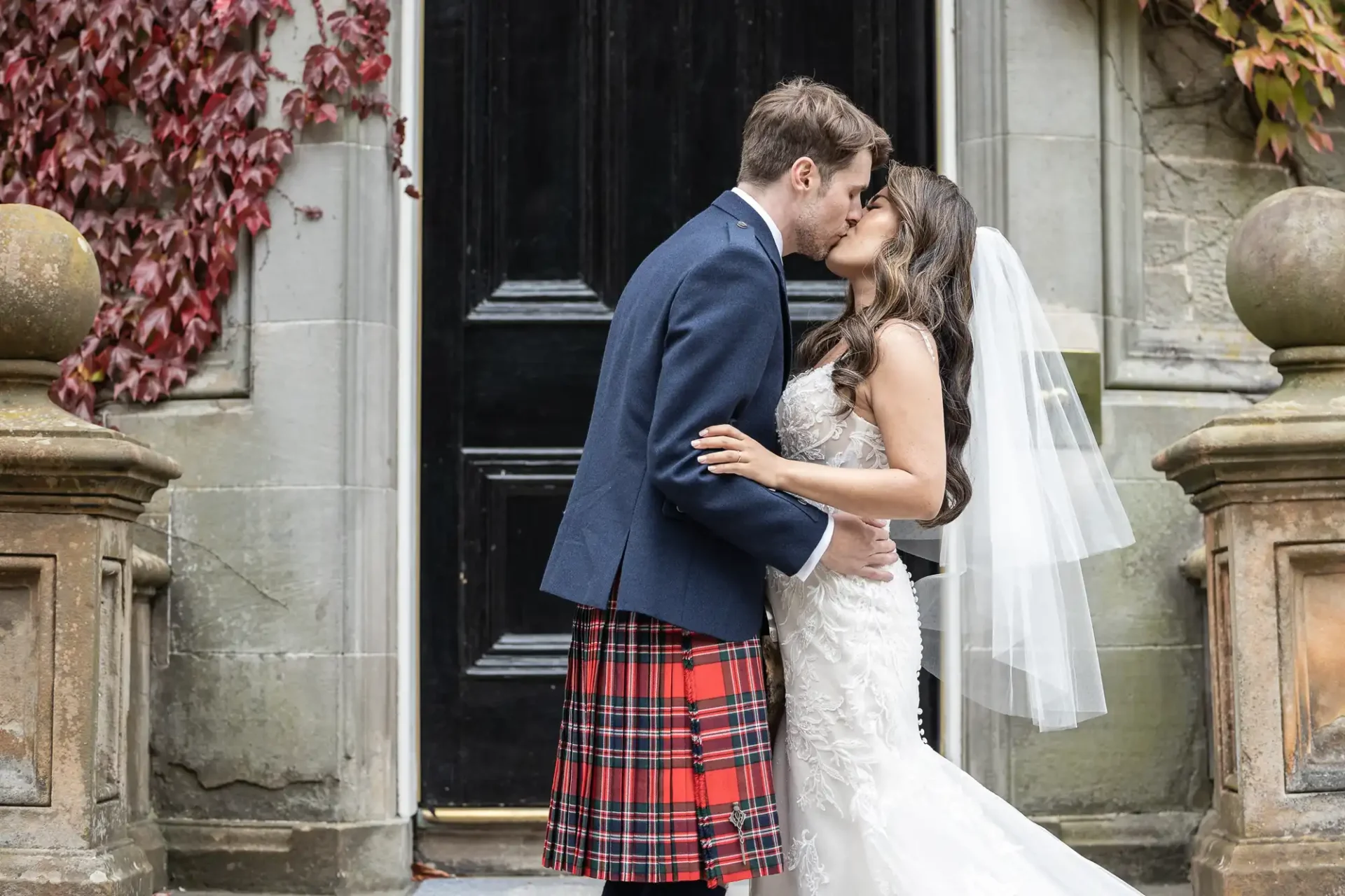 A bride and groom kiss in front of a dark door. The groom wears a tartan kilt and jacket; the bride wears a white dress and veil. Stone columns and red ivy frame the scene.