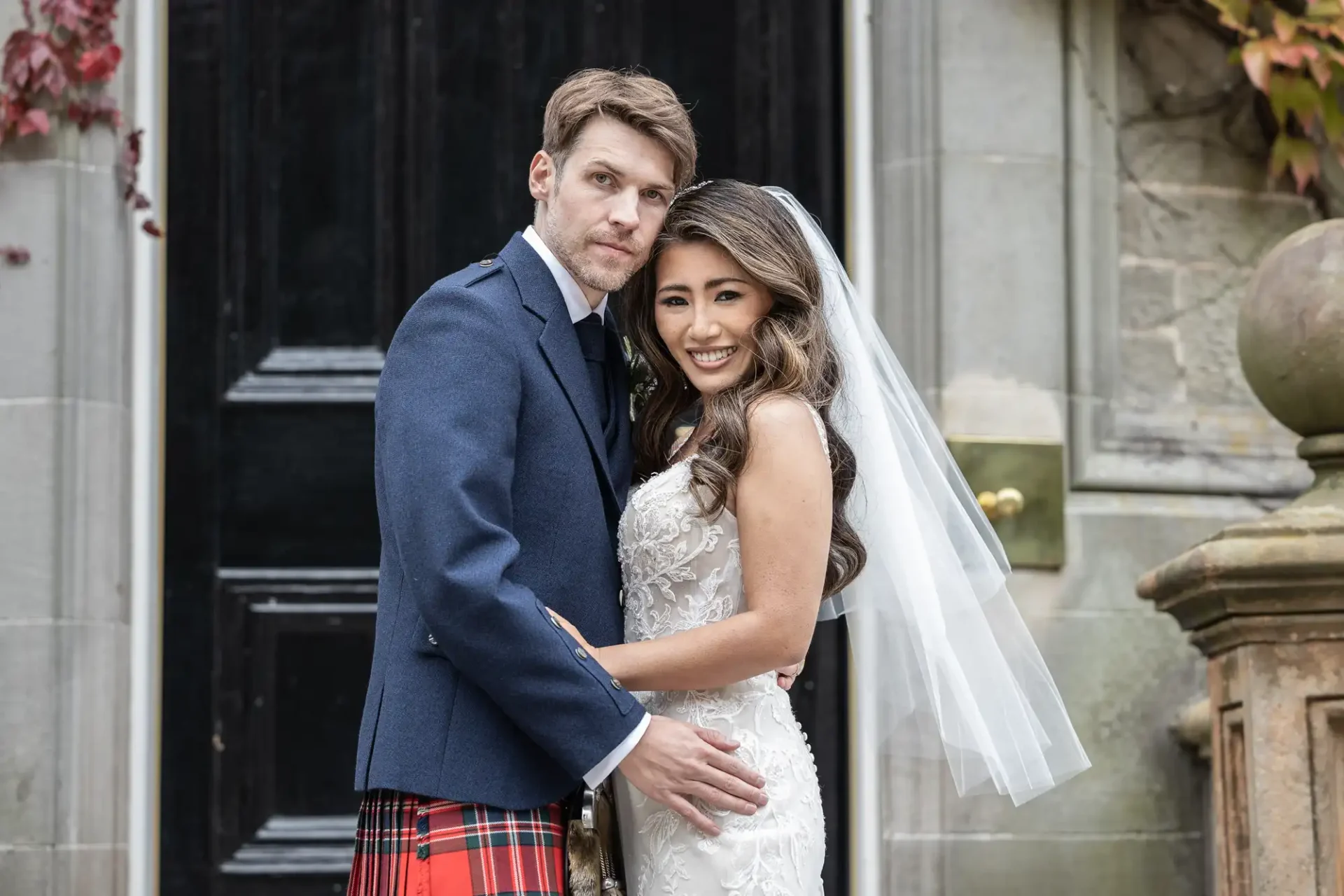 A bride in a lace gown and veil stands with a groom in a blue jacket and tartan kilt, posing in front of a dark wooden door.