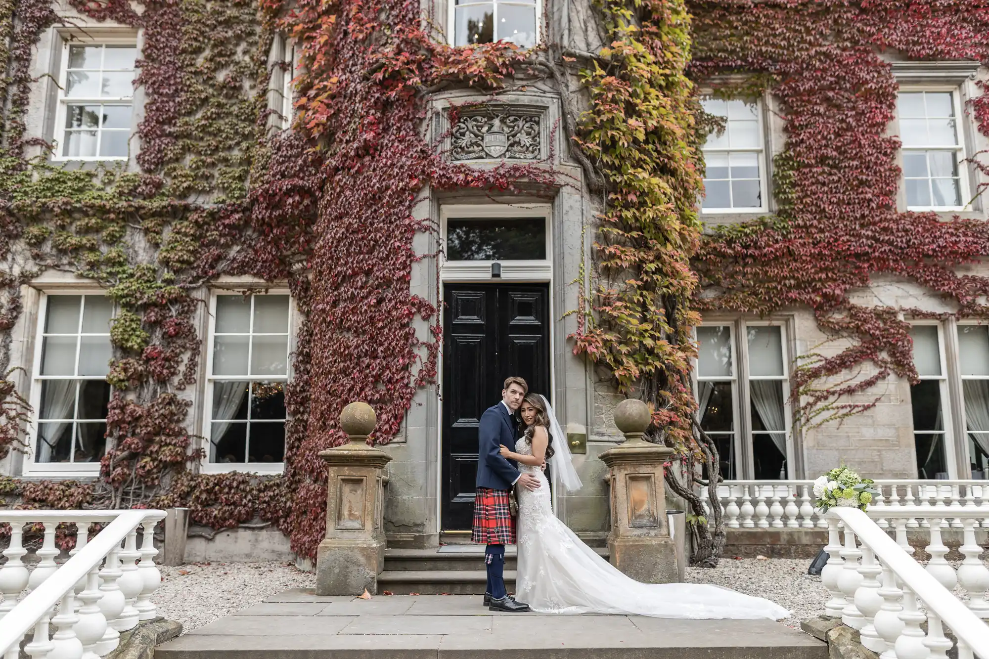 A couple in wedding attire stands embraced in front of an ivy-covered building with large windows and a black door. The groom wears a kilt, and the bride is in a long white dress with a train.