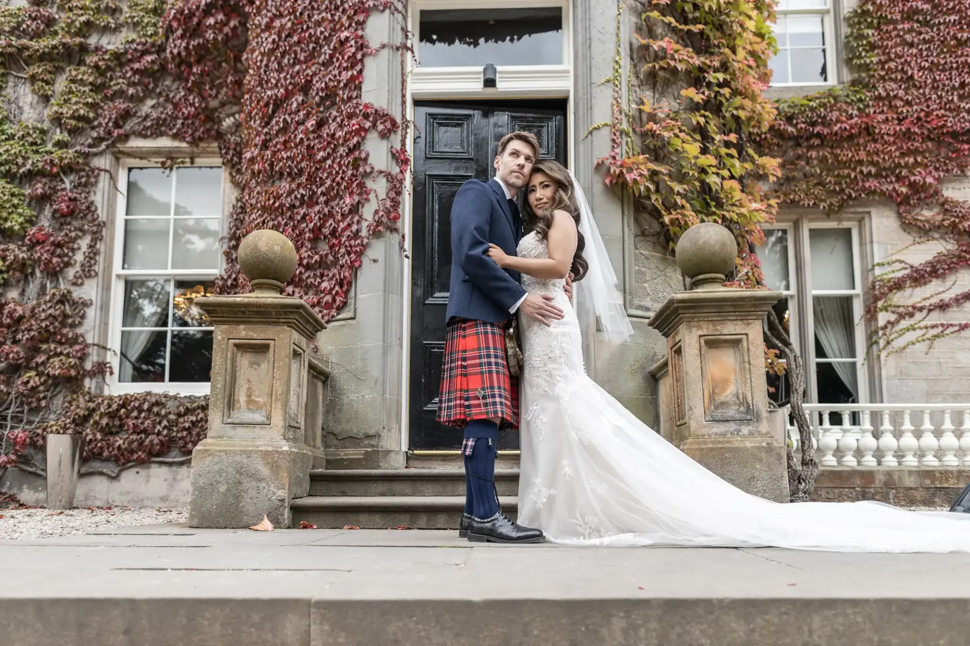 A couple poses on stone steps in front of a vine-covered building. The groom wears a Scottish kilt, and the bride is in a white dress with a long train.