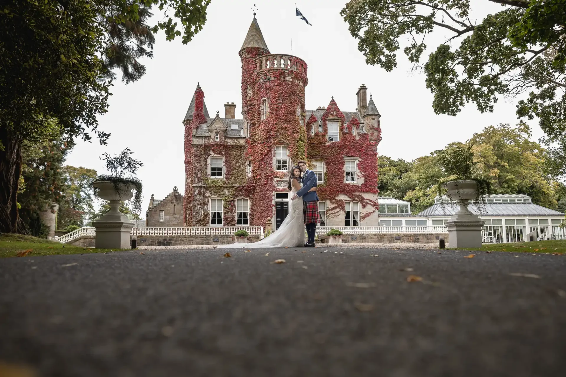 A couple in wedding attire stands in front of a large ivy-covered castle with a tall tower, surrounded by trees.