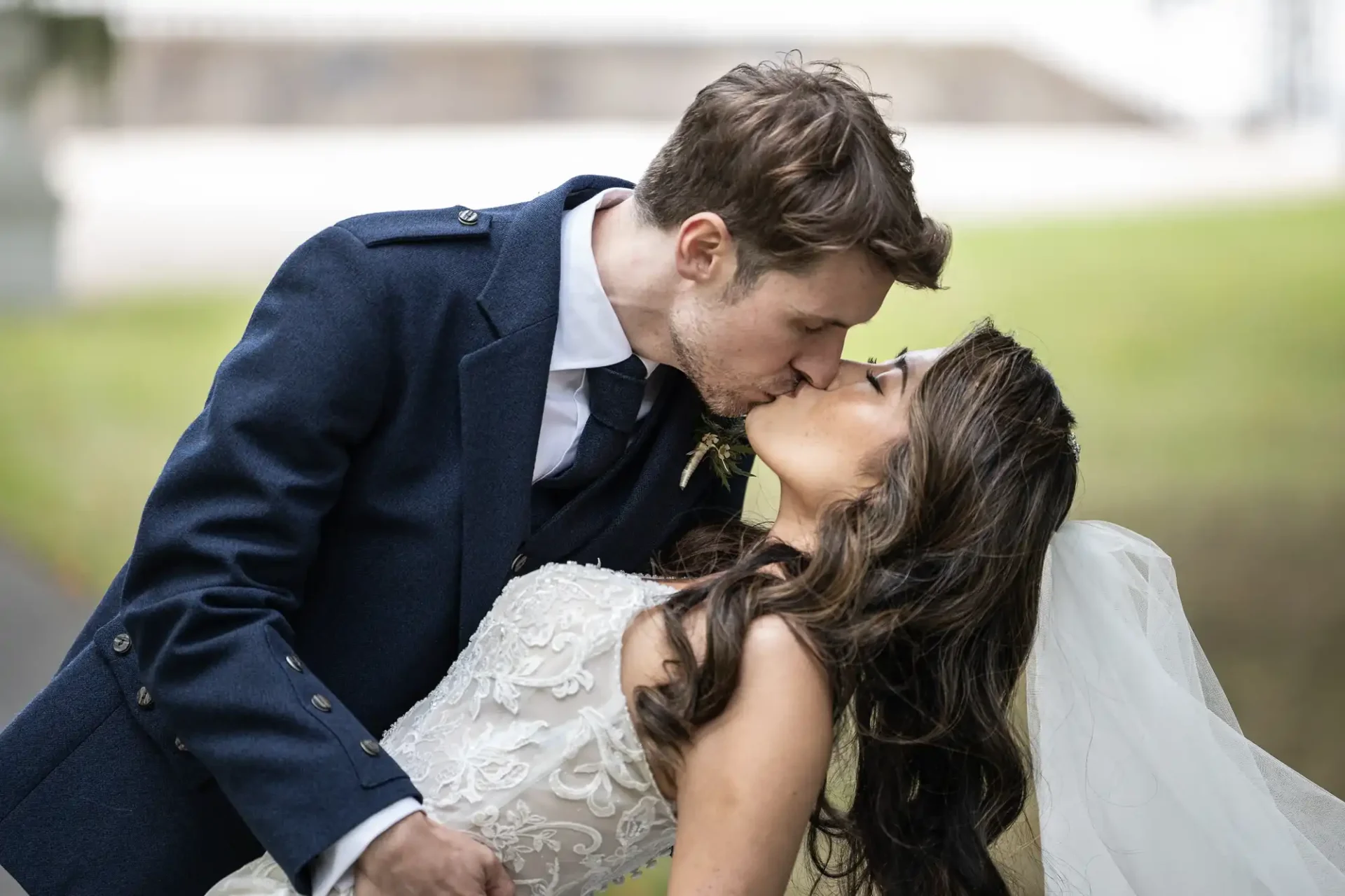 A bride and groom kiss outdoors. The groom is wearing a blue suit and the bride is in a white dress with a veil.