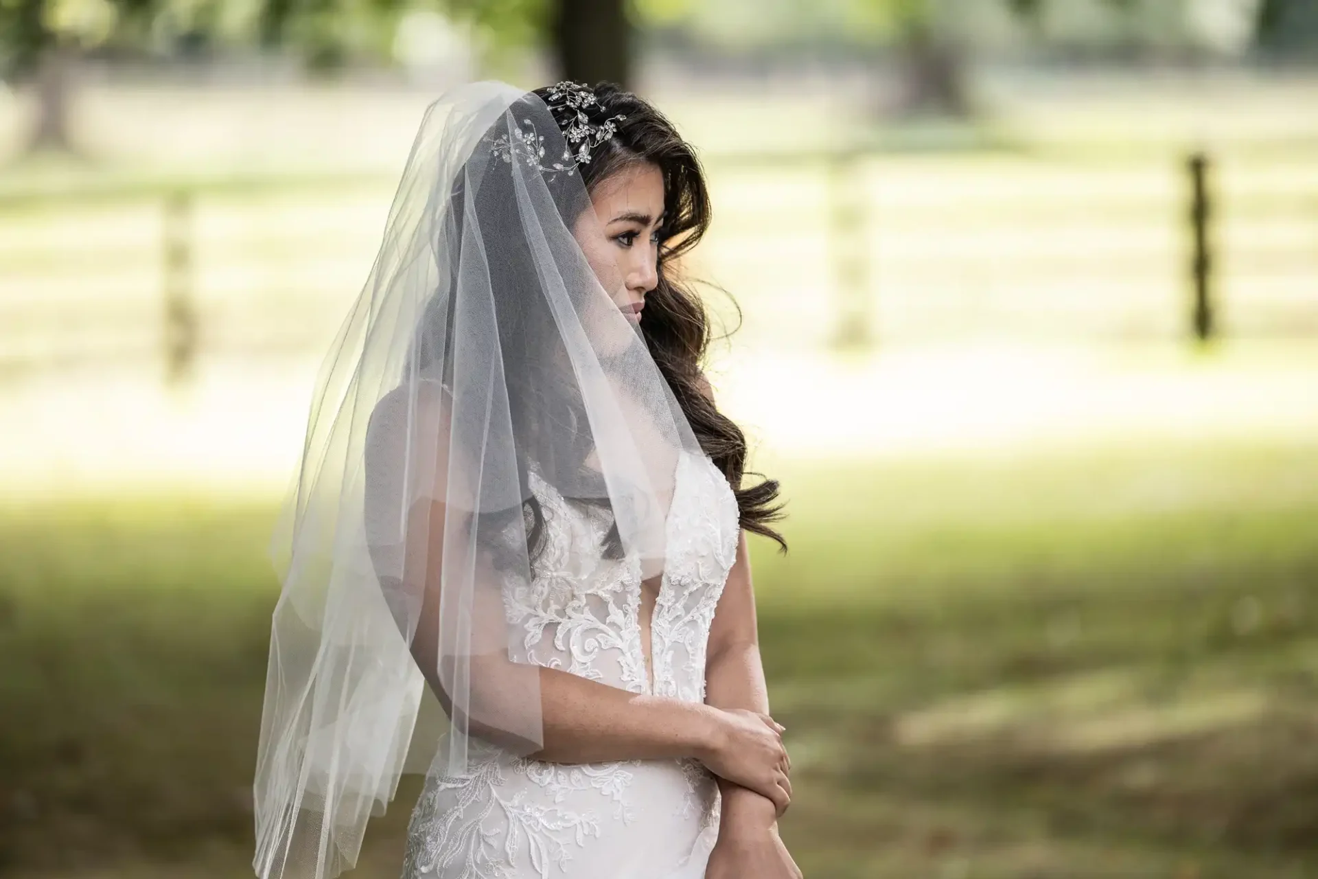 Bride in a white lace dress and veil stands outdoors, hands clasped, gazing to the side. Trees and a wooden fence are in the blurred background.