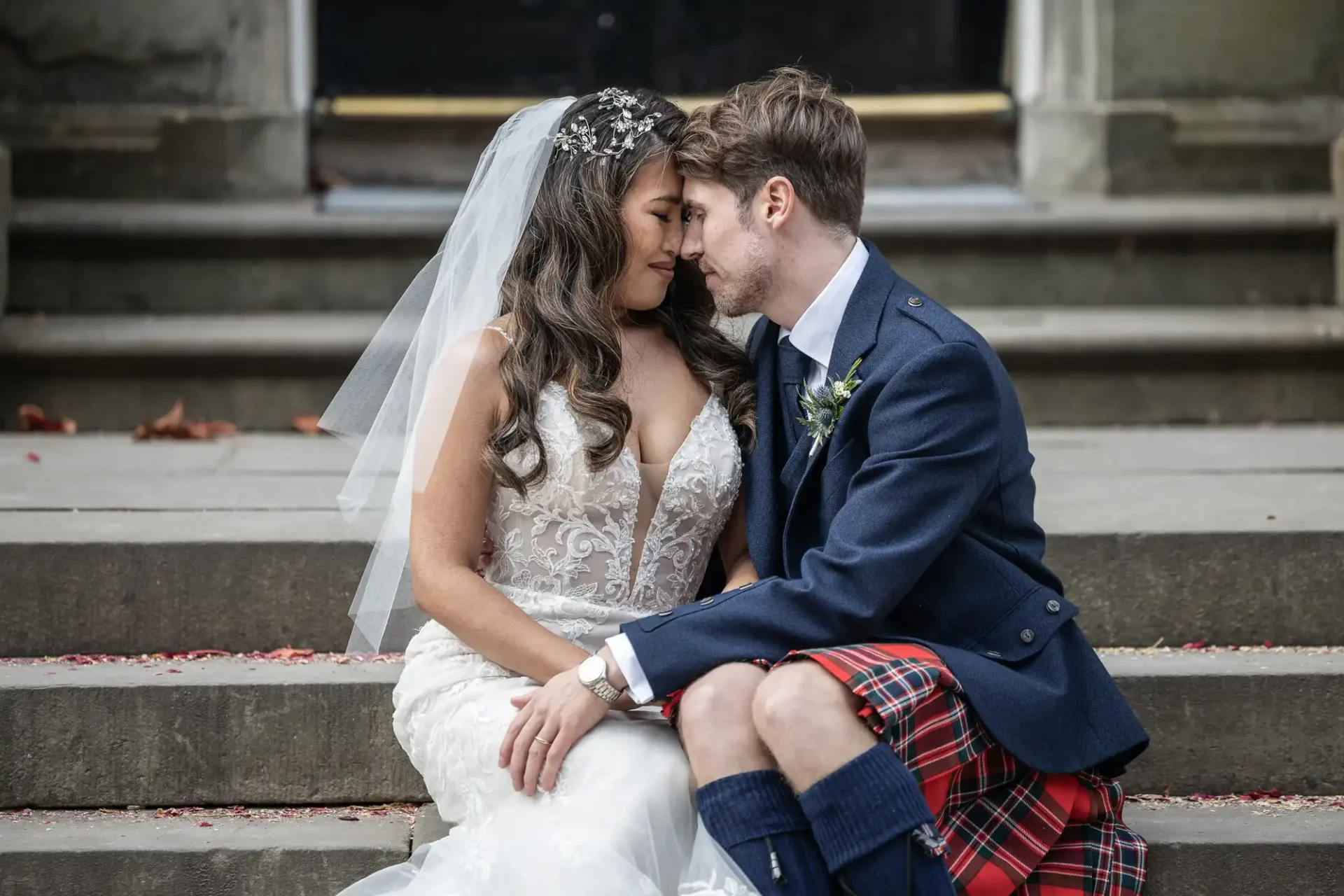 Bride in a white gown and groom in a kilt sit closely on stone steps, gently touching foreheads and holding hands.