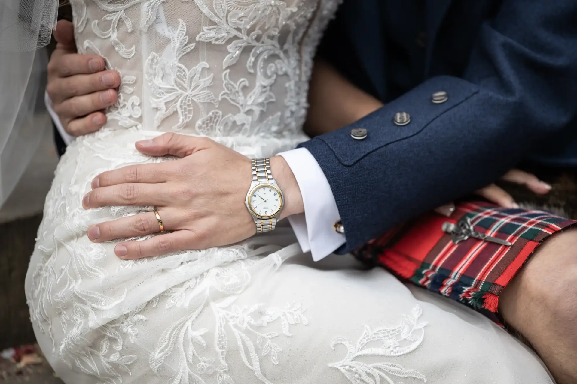 A close-up of a couple sitting, with the man's hand wearing a watch and wedding band on the woman's lace wedding dress. He's dressed in a kilt and a blue jacket with silver buttons.