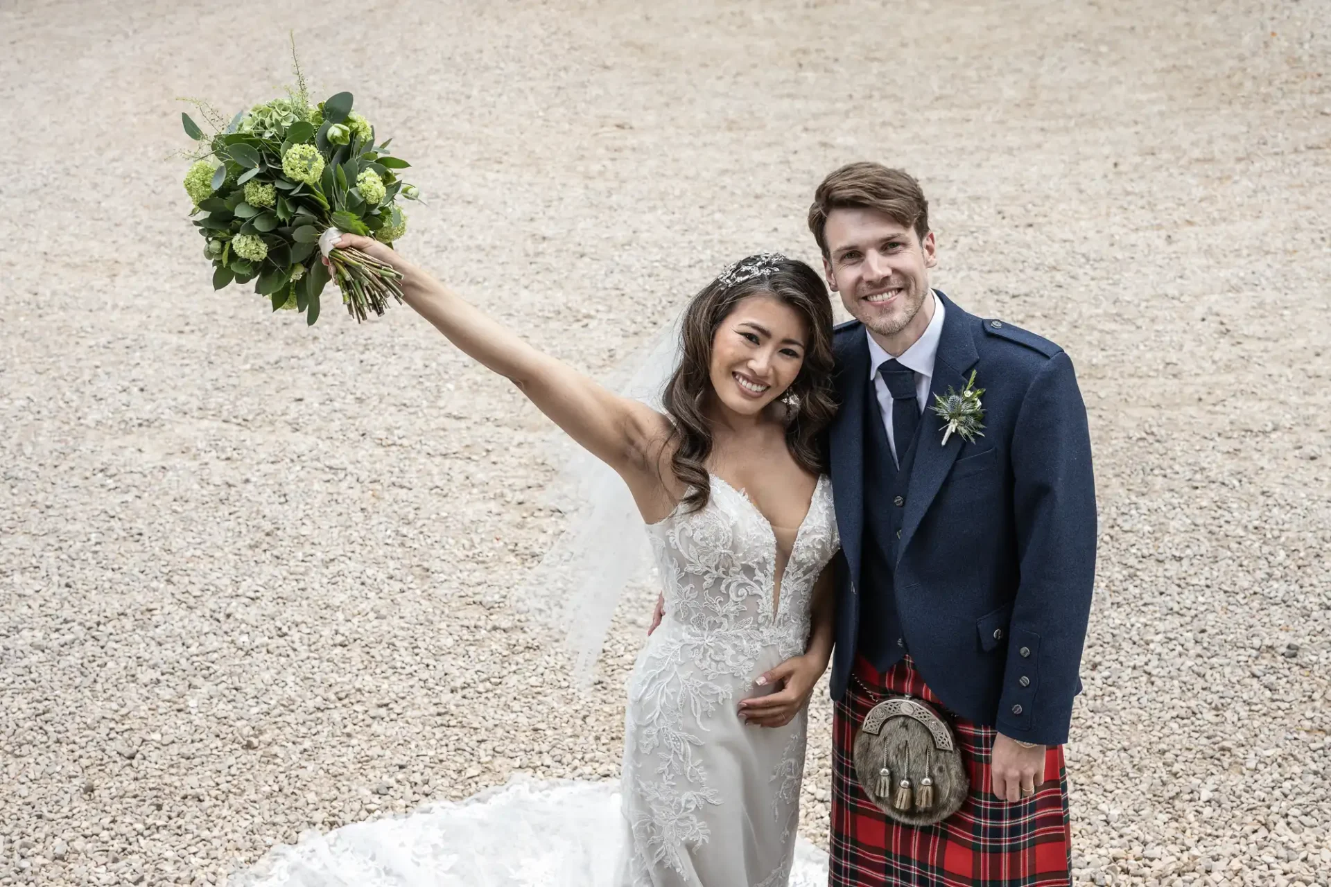 Bride holding a bouquet and groom wearing a kilt stand smiling on a gravel surface.