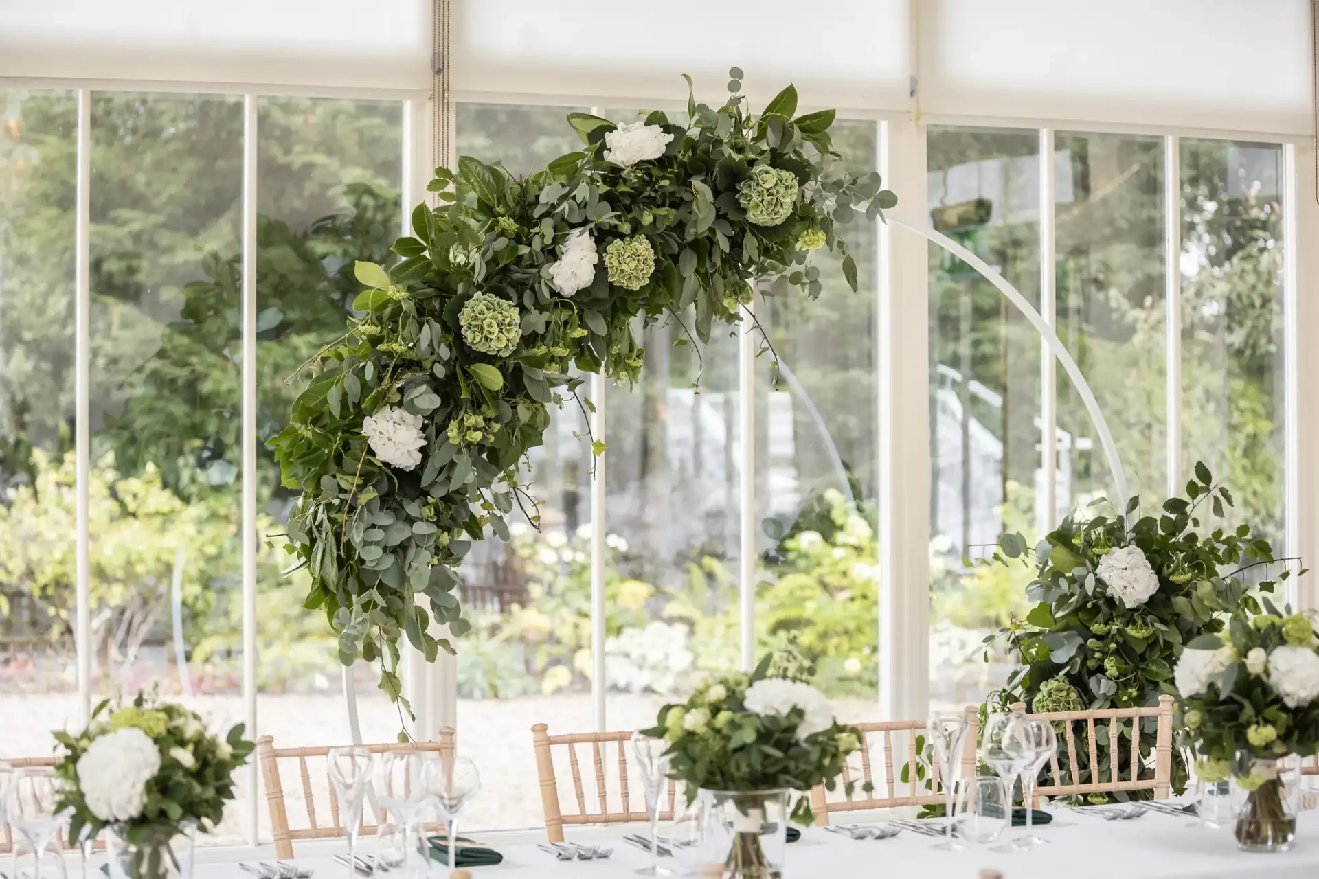 Elegant floral arrangement with white hydrangeas and greenery in a curved arch design above a table set with glasses and white napkins, in front of large windows.