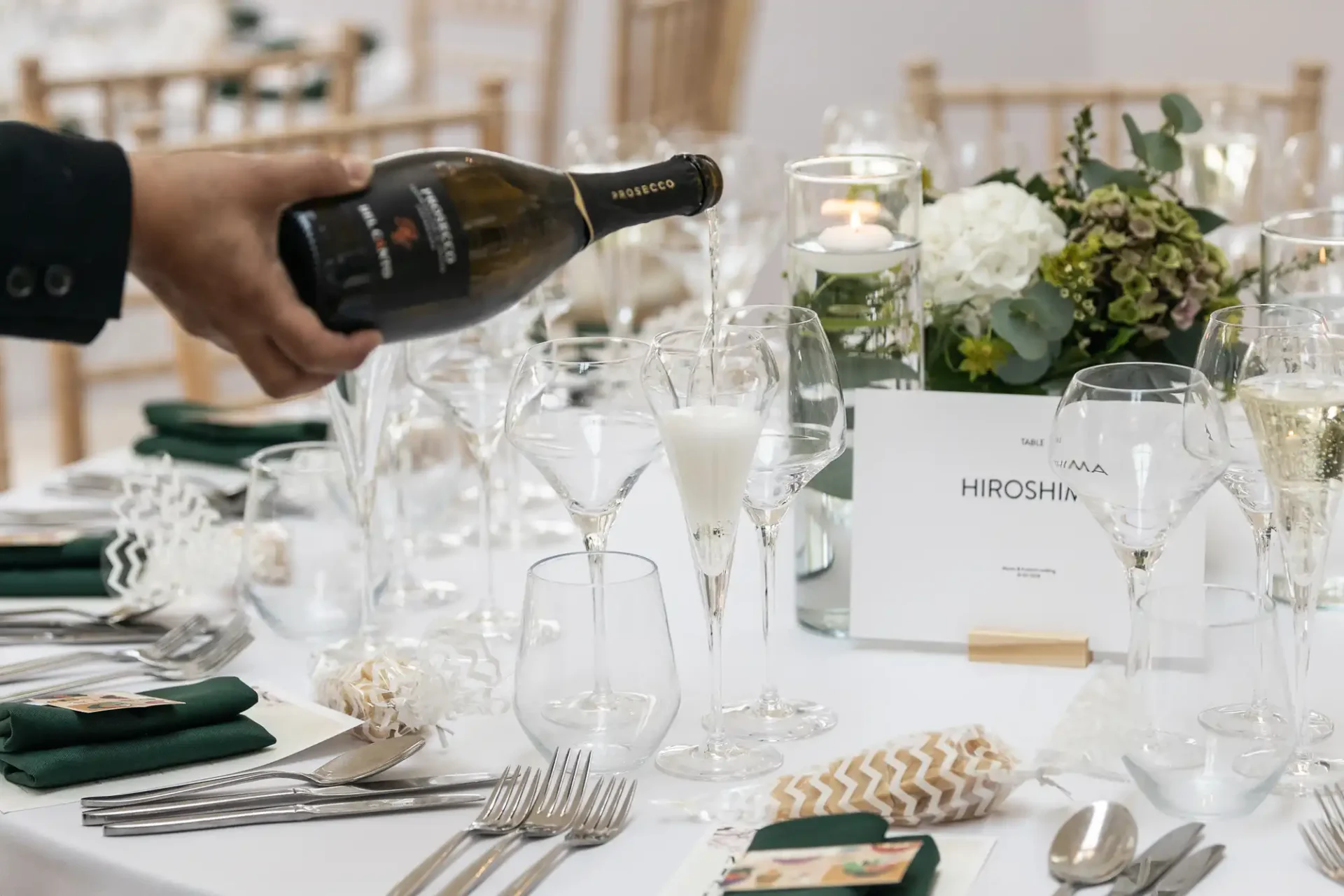 A hand pours champagne into a flute on a white-clothed table set with wine glasses, cutlery, and floral centerpieces. A card labeled "Hiroshima" is visible on the table.