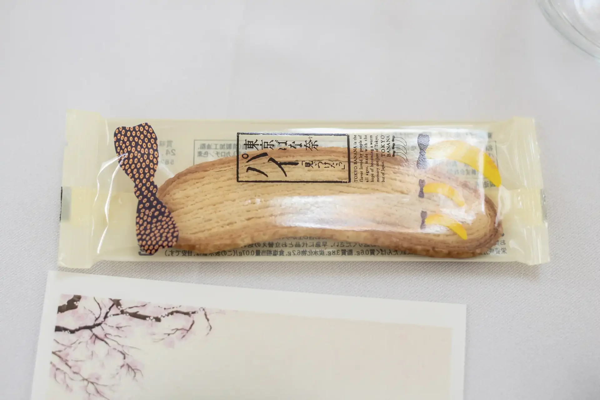 Packaged banana-shaped biscuit on a table next to a postcard with a cherry blossom design.