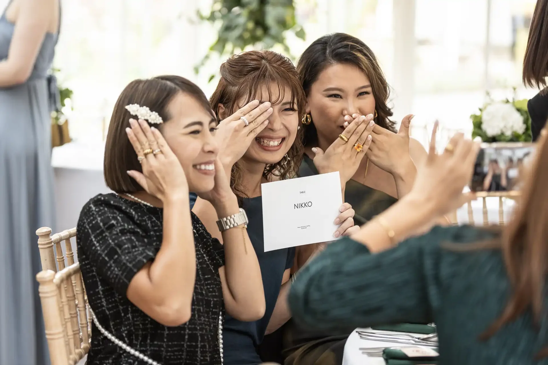 Three women are smiling and covering their faces playfully, sitting at a table, holding a card that says "NIKKO.