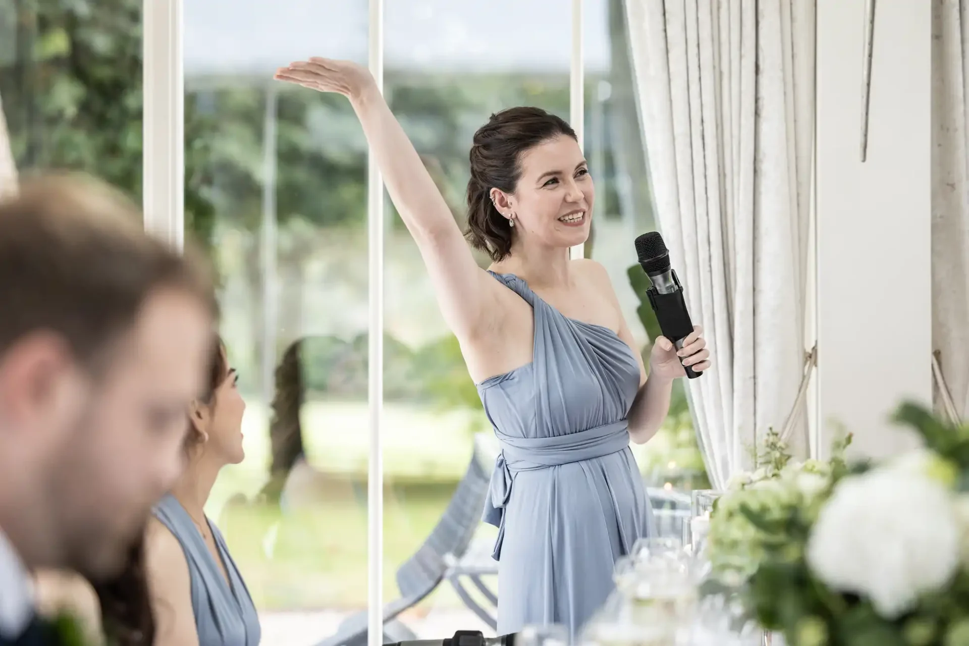 A woman in a blue dress holds a microphone and raises her left arm while speaking at an indoor event.