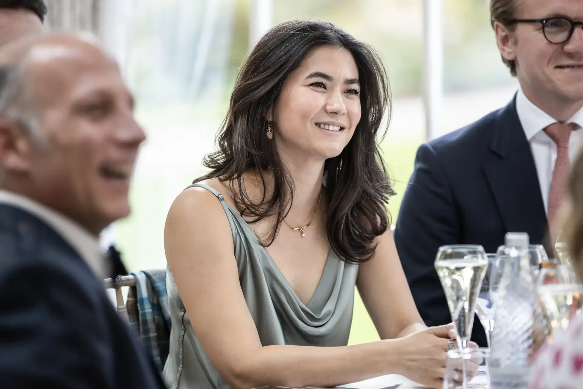 A woman smiling at a table, wearing a gray sleeveless top, with champagne glasses and two people in the background.