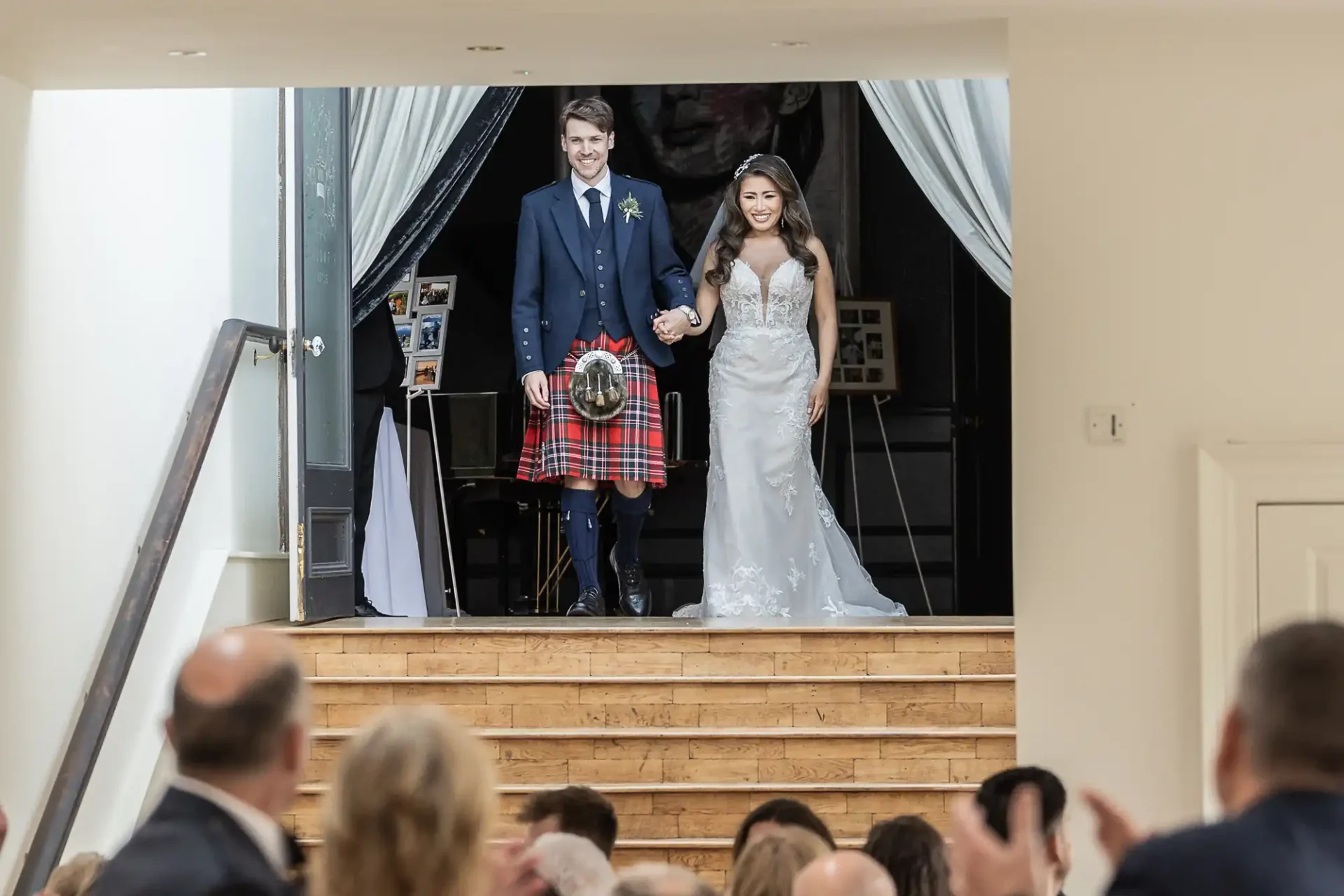 A bride and groom hold hands and smile at the top of a staircase, dressed in a wedding gown and kilt, as guests look on from below.