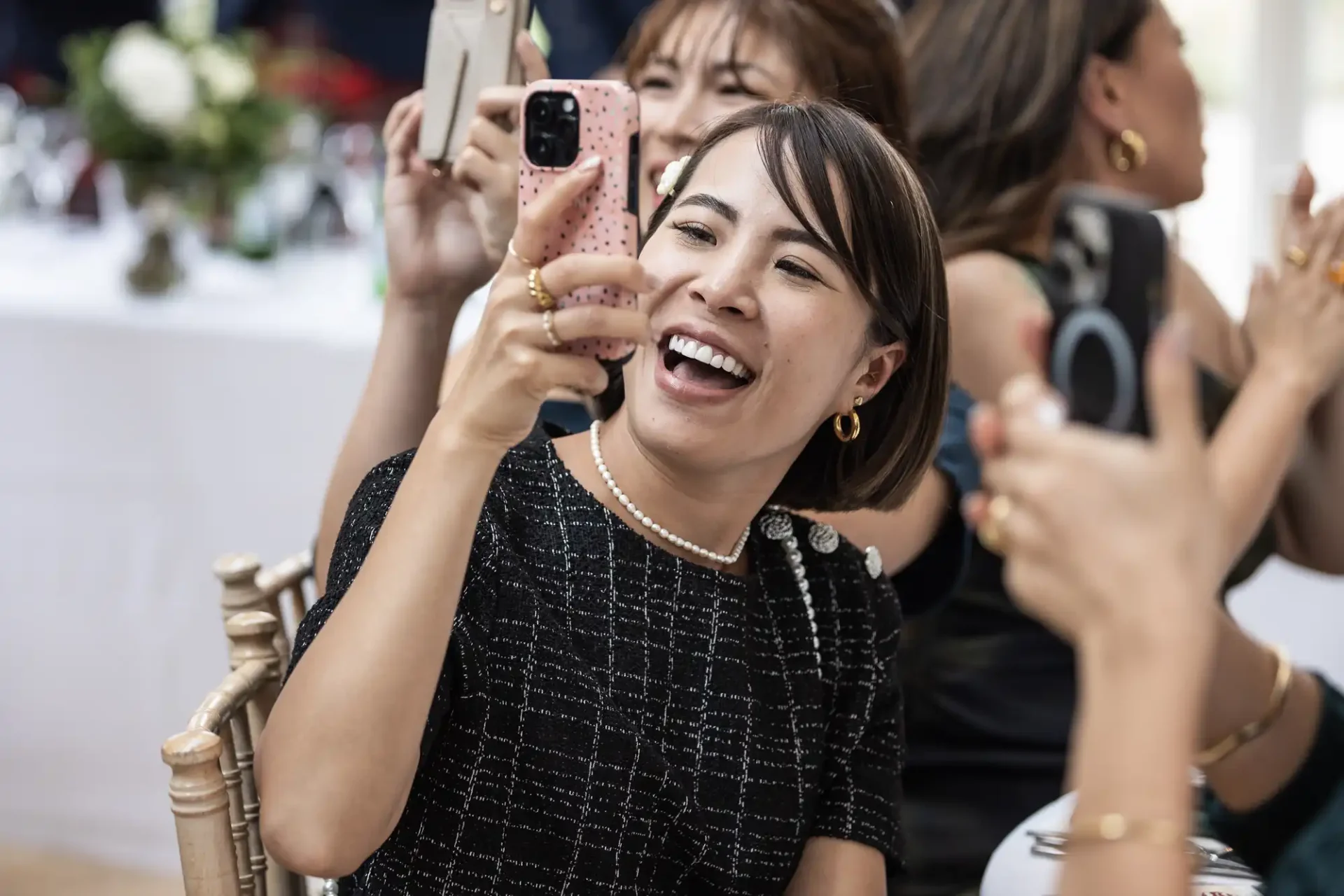 A woman in a black dress with gold jewelry smiles while taking a photo with her smartphone at a social gathering. Other people and tables are visible in the background.