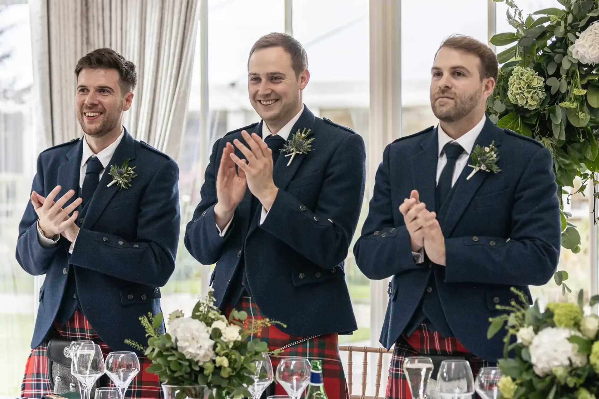 Three men in matching blue jackets and red tartan kilts clap and smile at an indoor event.