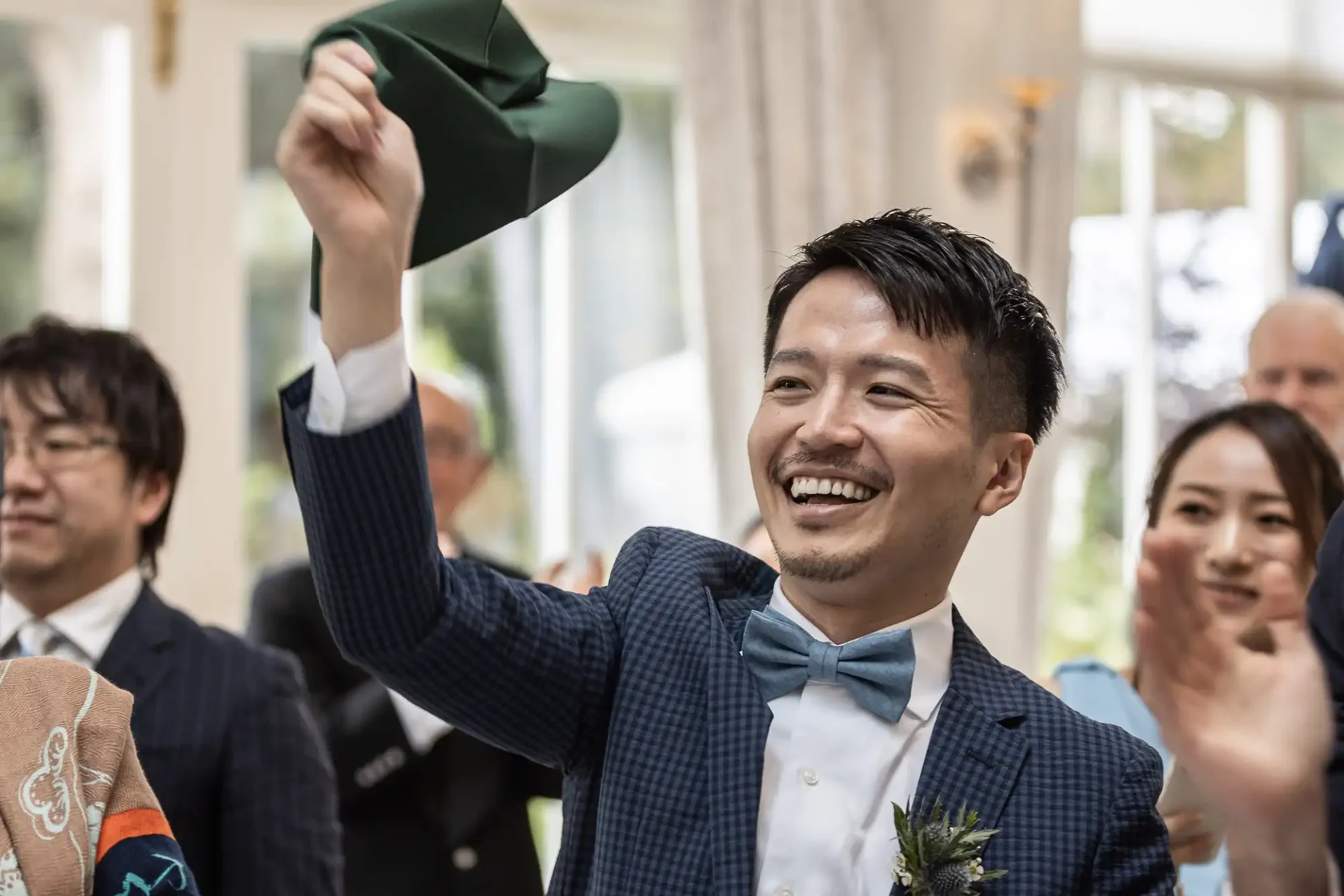 A man in a blue suit and bow tie smiles while holding up a green hat. People are in the background, and the setting appears to be a celebratory event.