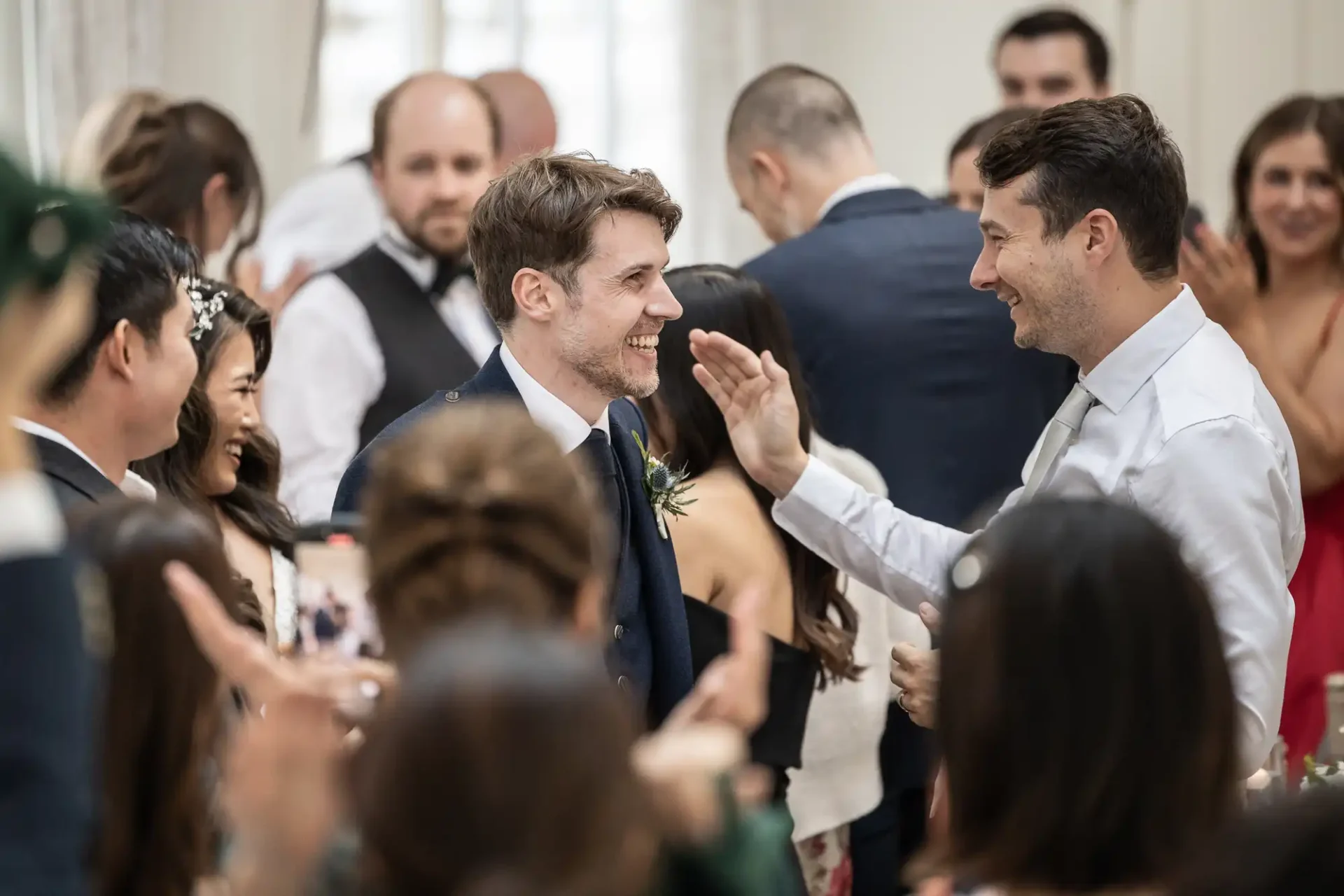 A groom in a blue suit smiles as he is congratulated by guests during a wedding reception.