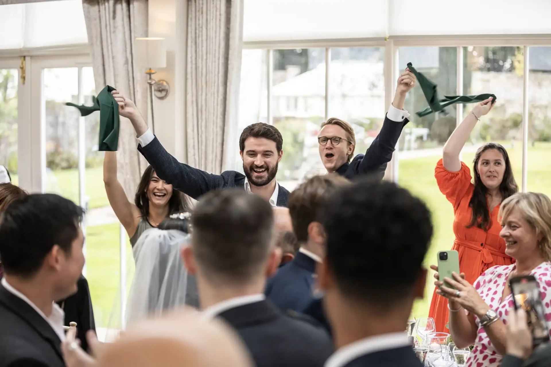 People in formal attire cheer and hold up green cloths at an indoor event with large windows in the background.