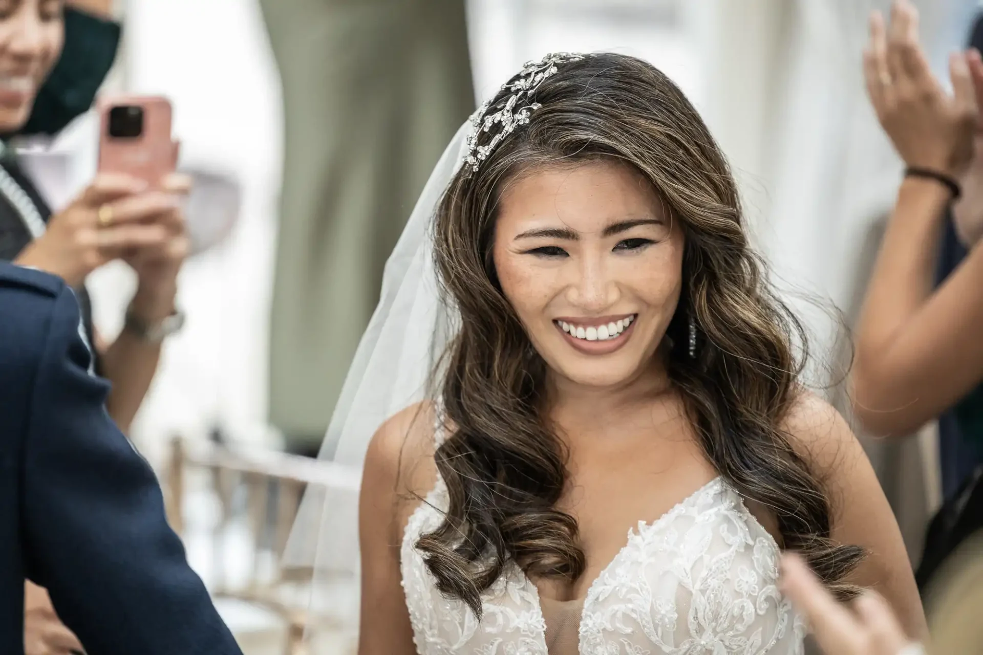 A bride with long, wavy hair and a detailed white gown smiles as people around her take photos with phones.