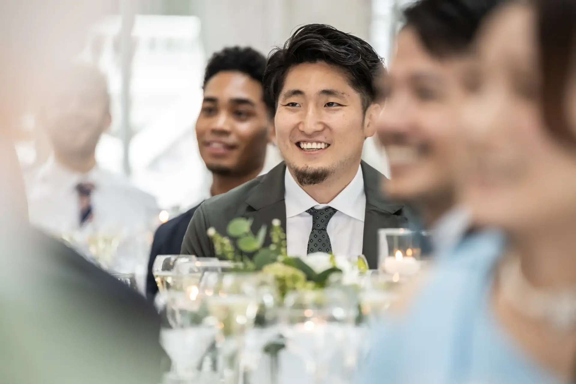 Man in suit smiling, sitting among others at a formal event with tables decorated with flowers and candles.