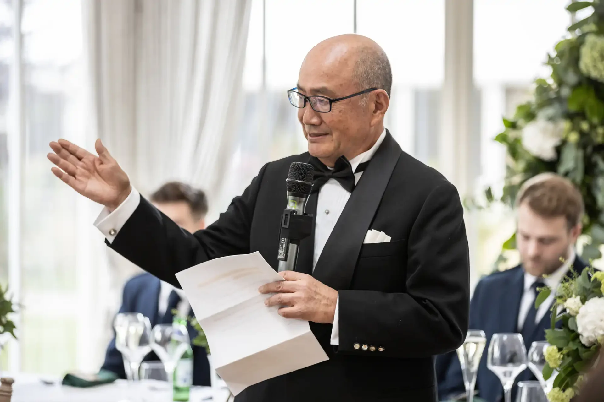 A man in a tuxedo speaks into a microphone, holding a paper. He gestures with his other hand. There is a floral arrangement and people blurred in the background.