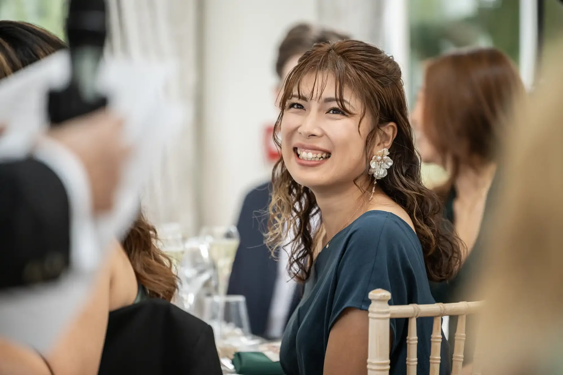 Woman in blue dress smiling and sitting at a formal event, with blurred people and tables in the background.