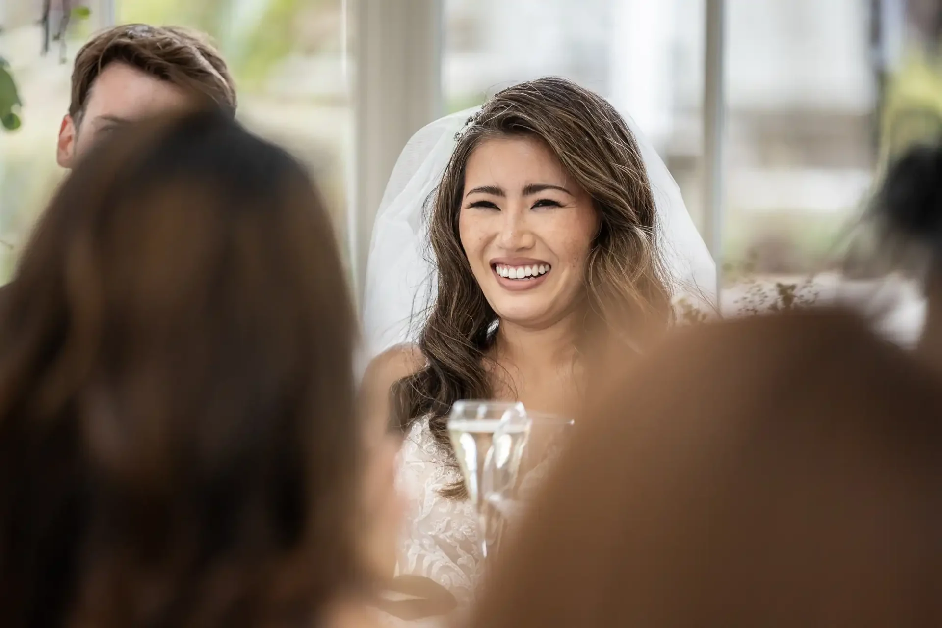 A bride with a veil smiles while sitting indoors, surrounded by people at a gathering.